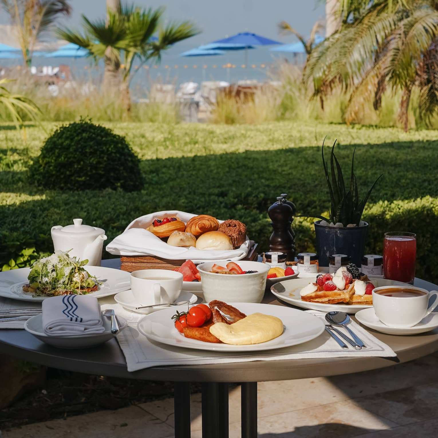 Outoor table filled with plates and bowls of breakfast food with a green lawn nearby and the ocean in the distance