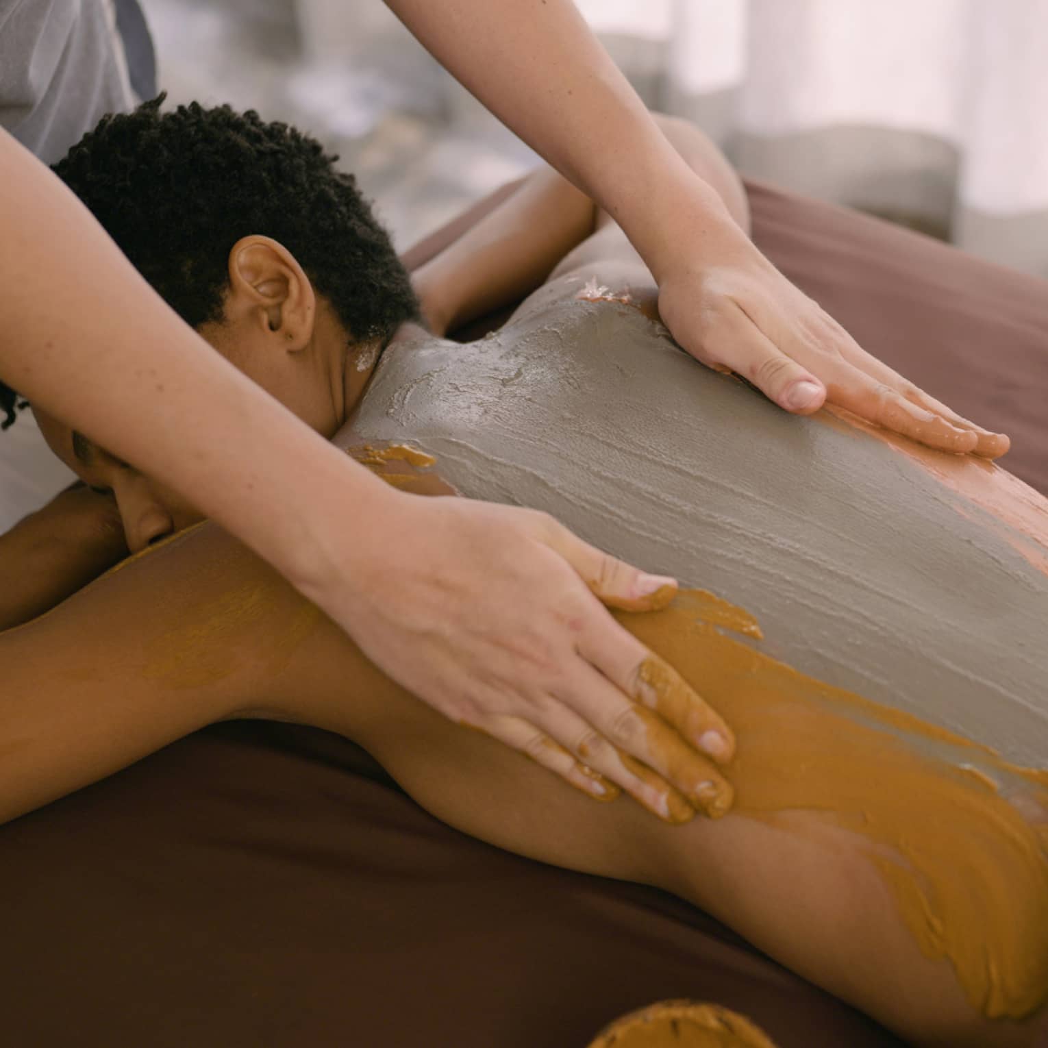 Hands rub clay across person's back as they lay on massage table