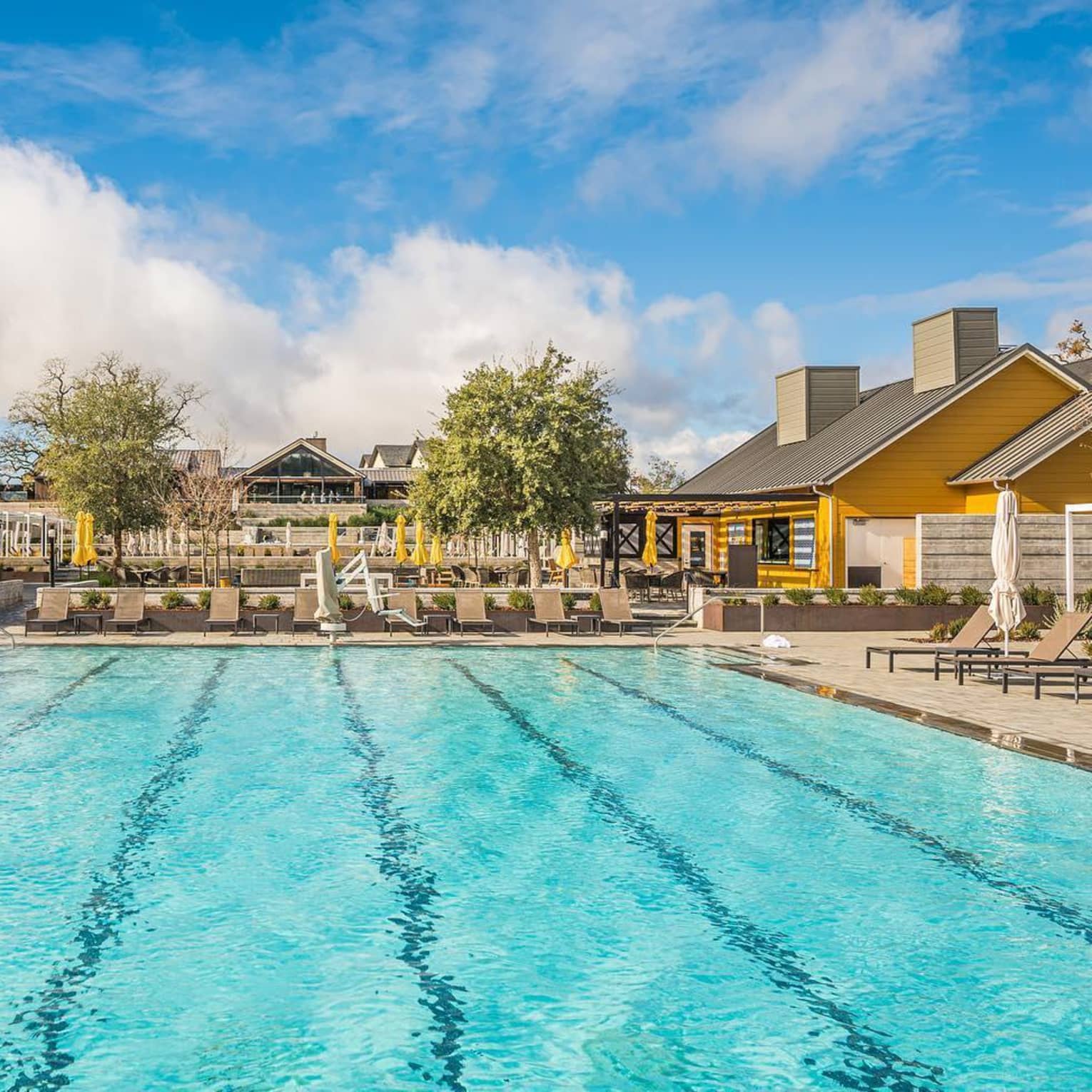 Outdoor swimming pool with lounge chairs and umbrellas, surrounded by buildings and trees under a partly cloudy sky.