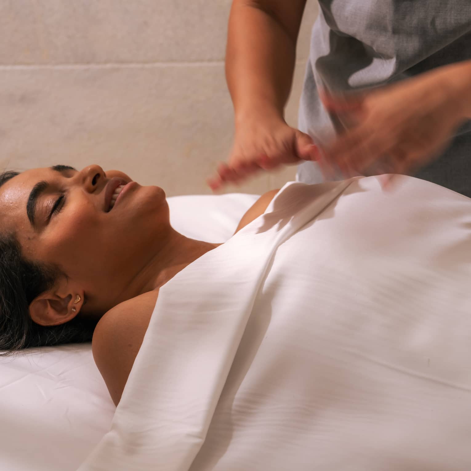 Person lies beneath a white sheet with head and shoulders shown as another person hovers hands over their body