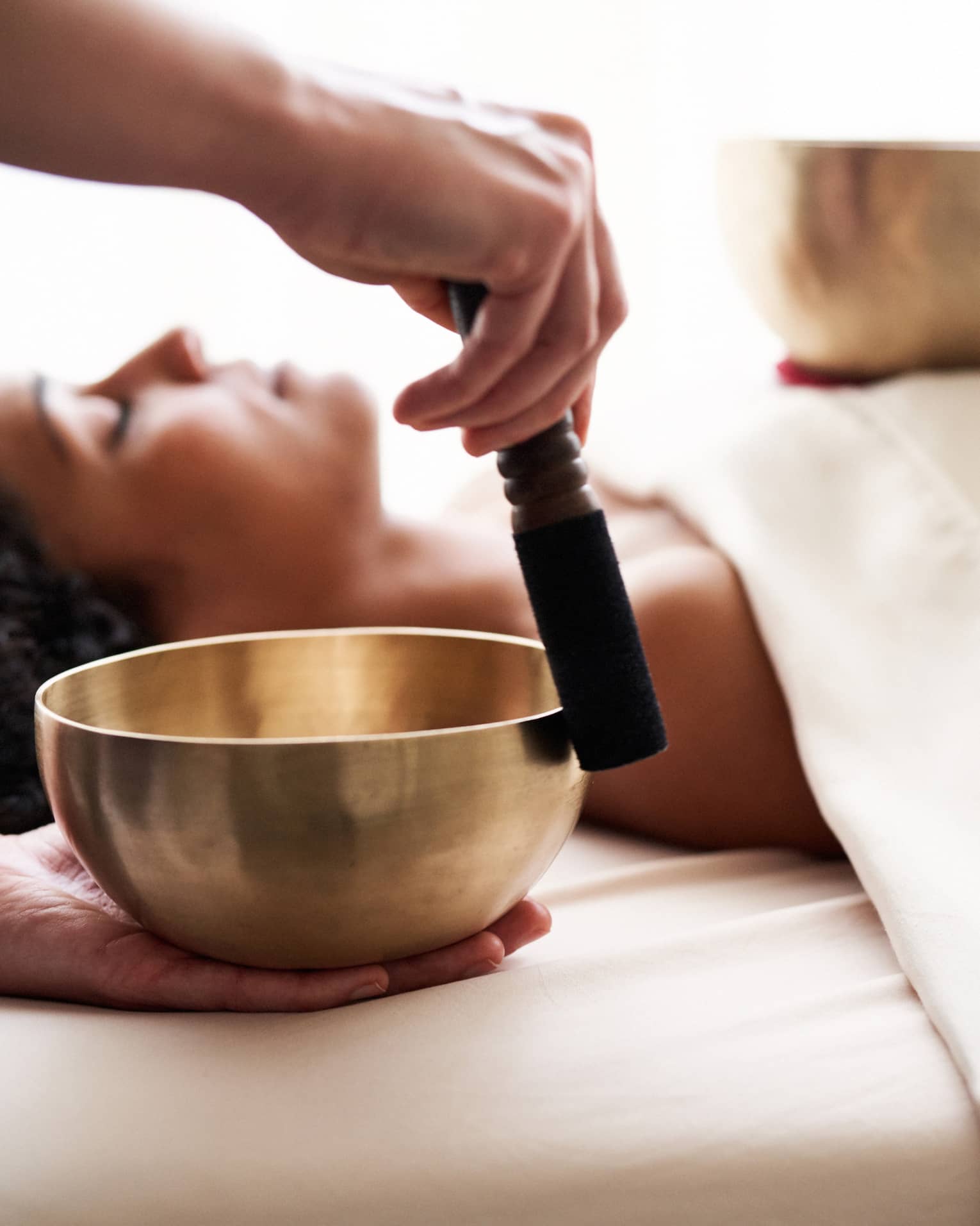 Woman with eyes closed lies on massage table in as vibrational sound therapy is performed with a Tibetan singing bowl