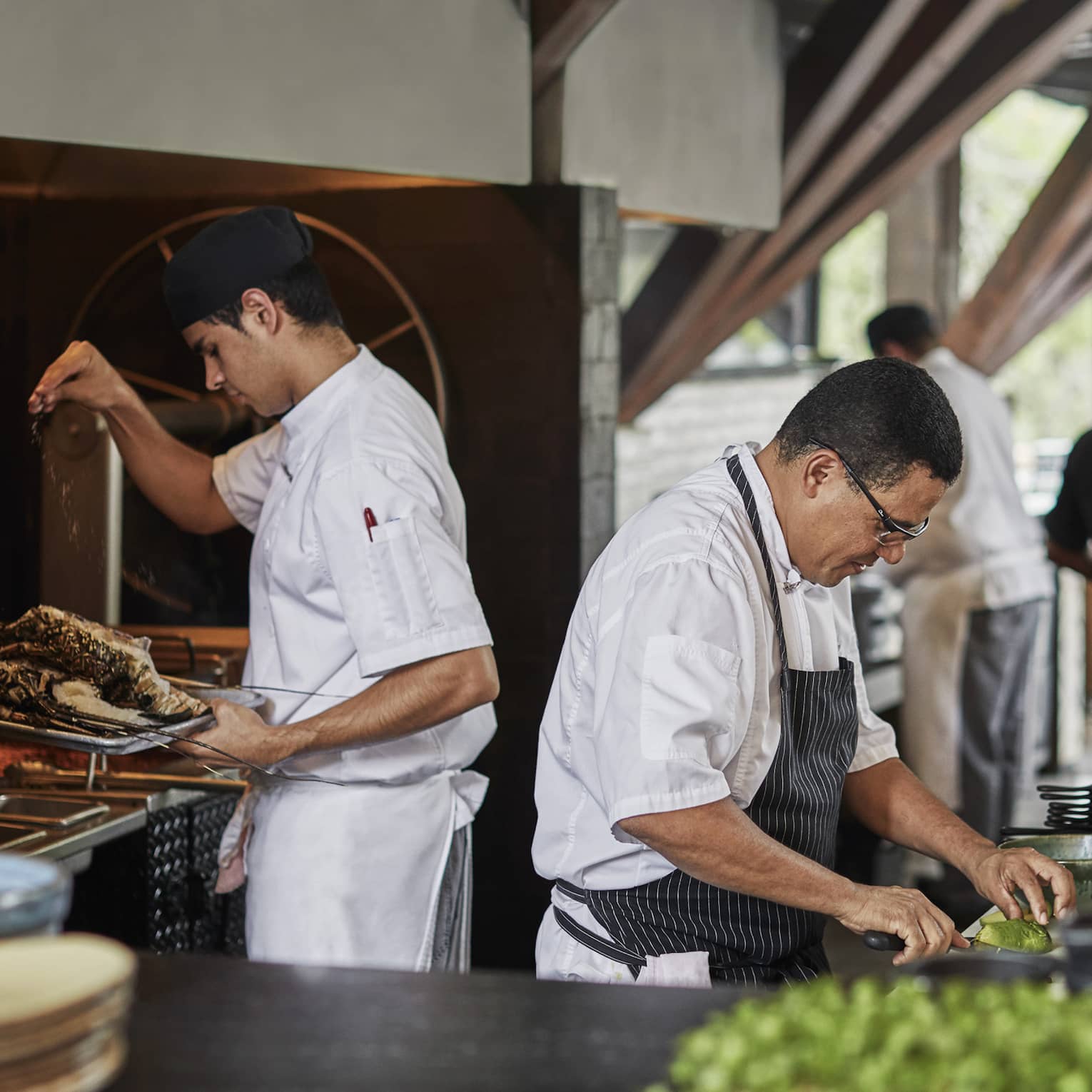As one chef seasons roast meat at the grill, another slices avocado in a sunny open kitchen with wooden ceiling beams.