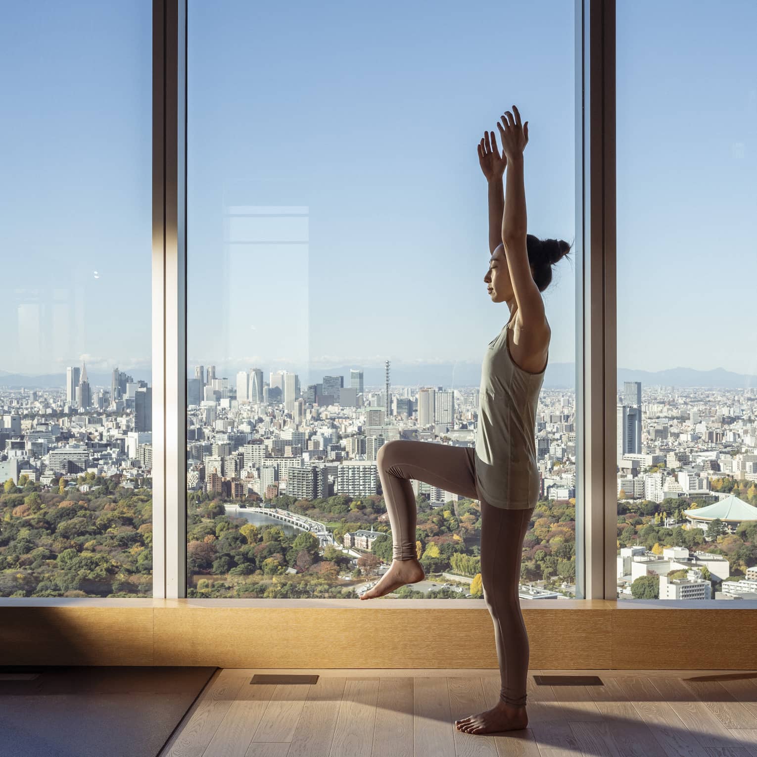Woman stands in yoga pose in room with floor-to-ceiling windows with city views