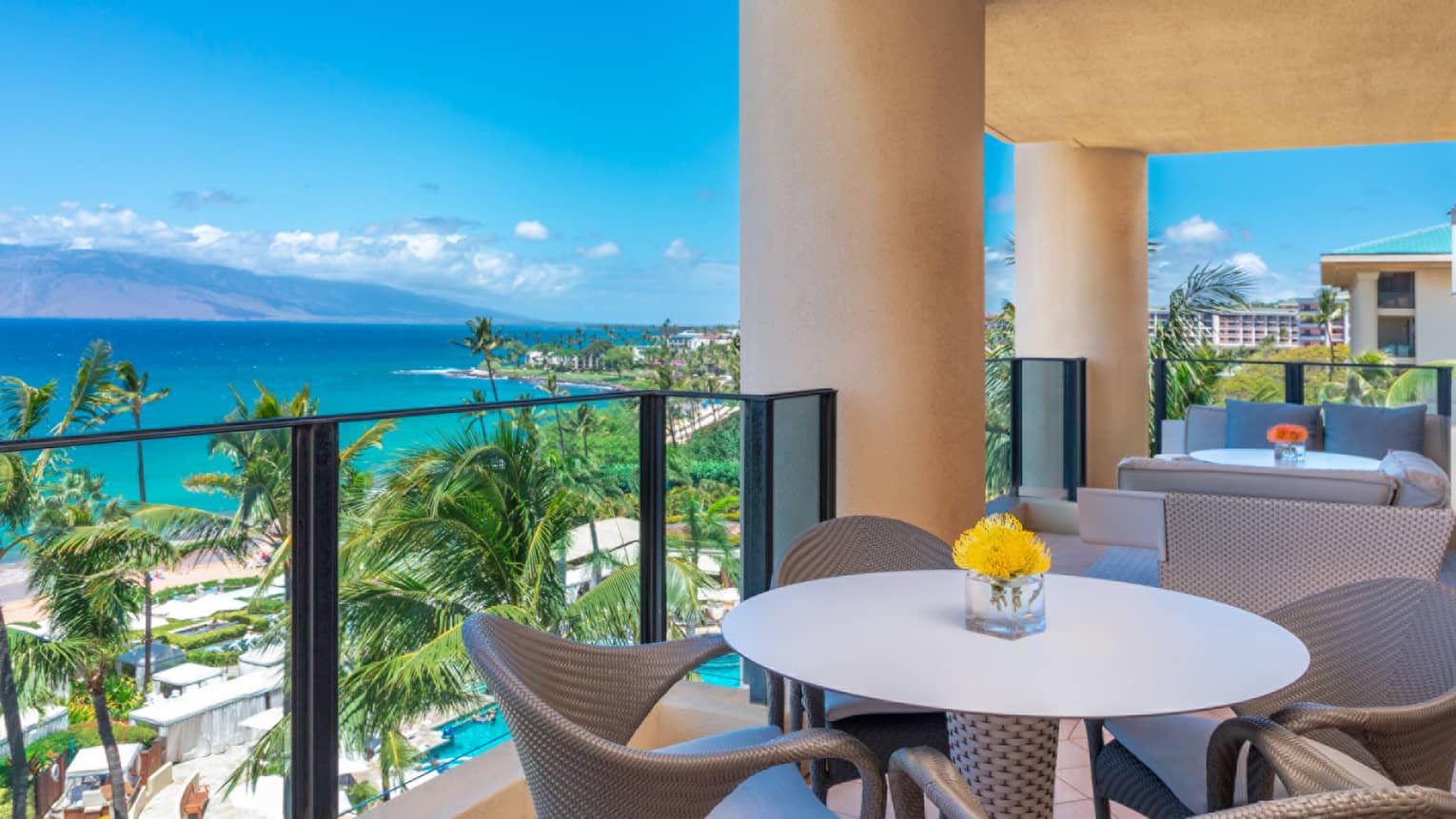 Balcony with round dining table, cushioned seating, ocean view and palm trees under a bright blue sky