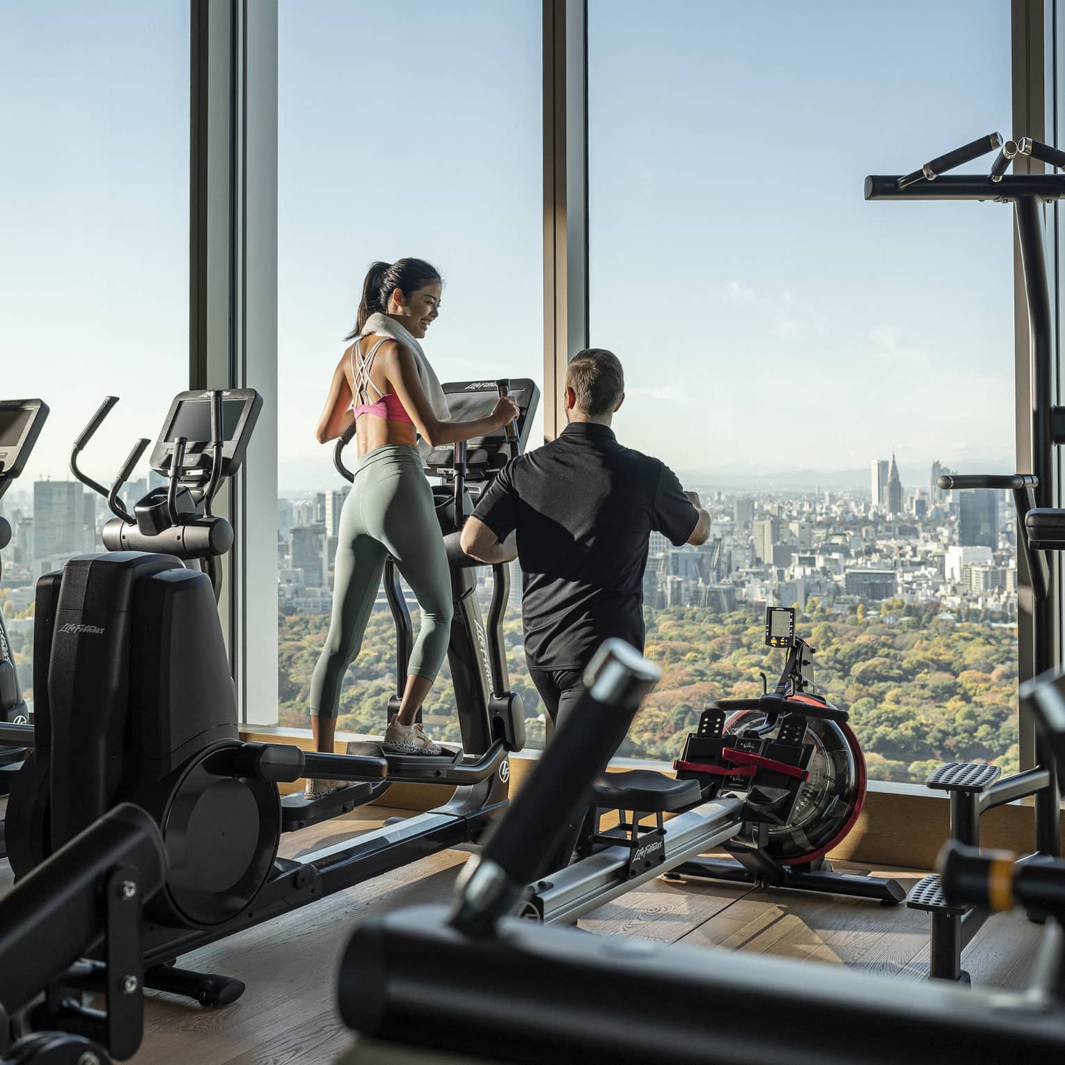 Woman trains on treadmill with male trainer, overlooking downtown Tokyo