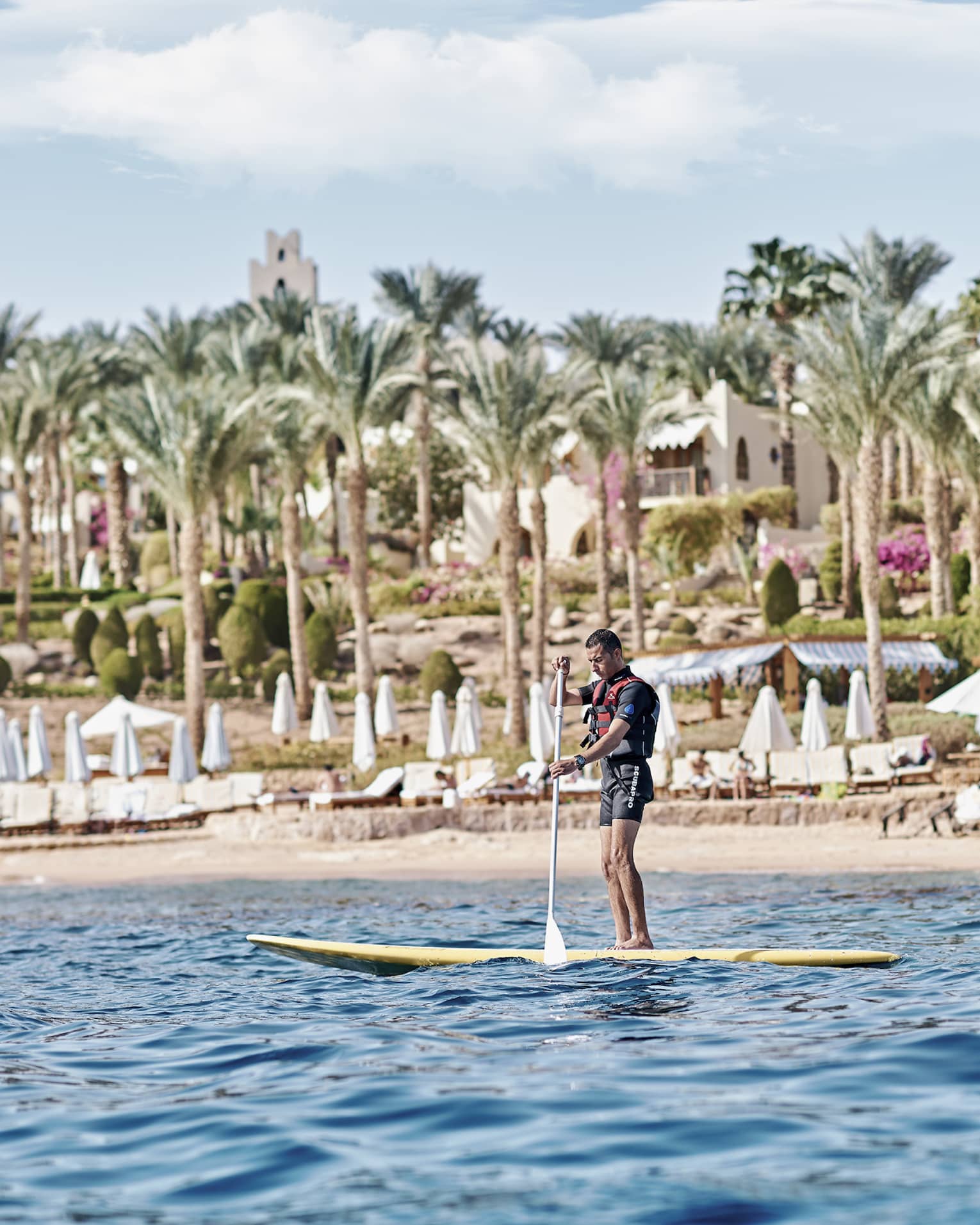 A paddler stands on a board in the water in front of a beach lined with lounge chairs and palm trees and a villa beyond.