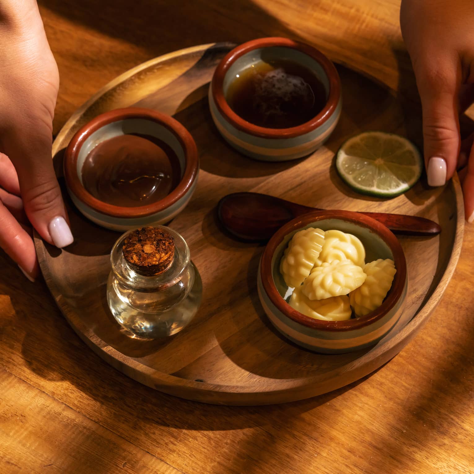 Pair of hands places a round wooden platter holding three small stoneware bowls each filled with spa ingredients, a small wooden spon, a small glass bottle of oil and a slice of lime
