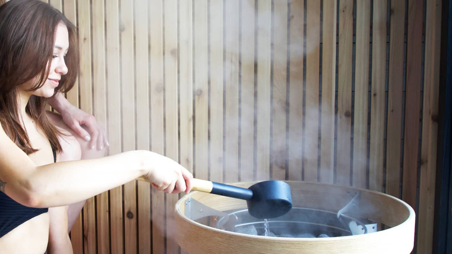 Two women pouring water onto hot rocks creating steam.