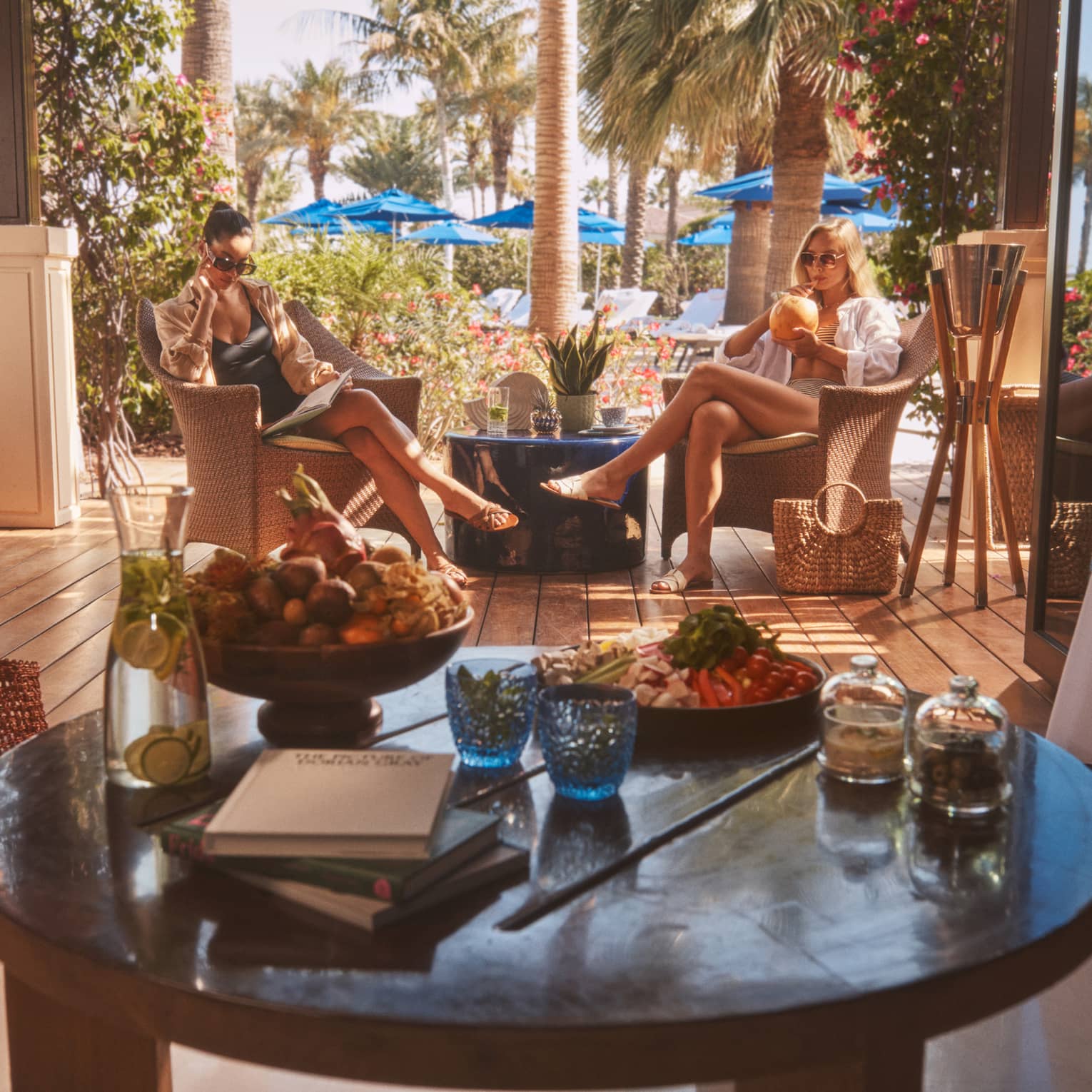 Two people sitting in rattan chairs with round table in forefront and blue pool umbrellas in backdrop