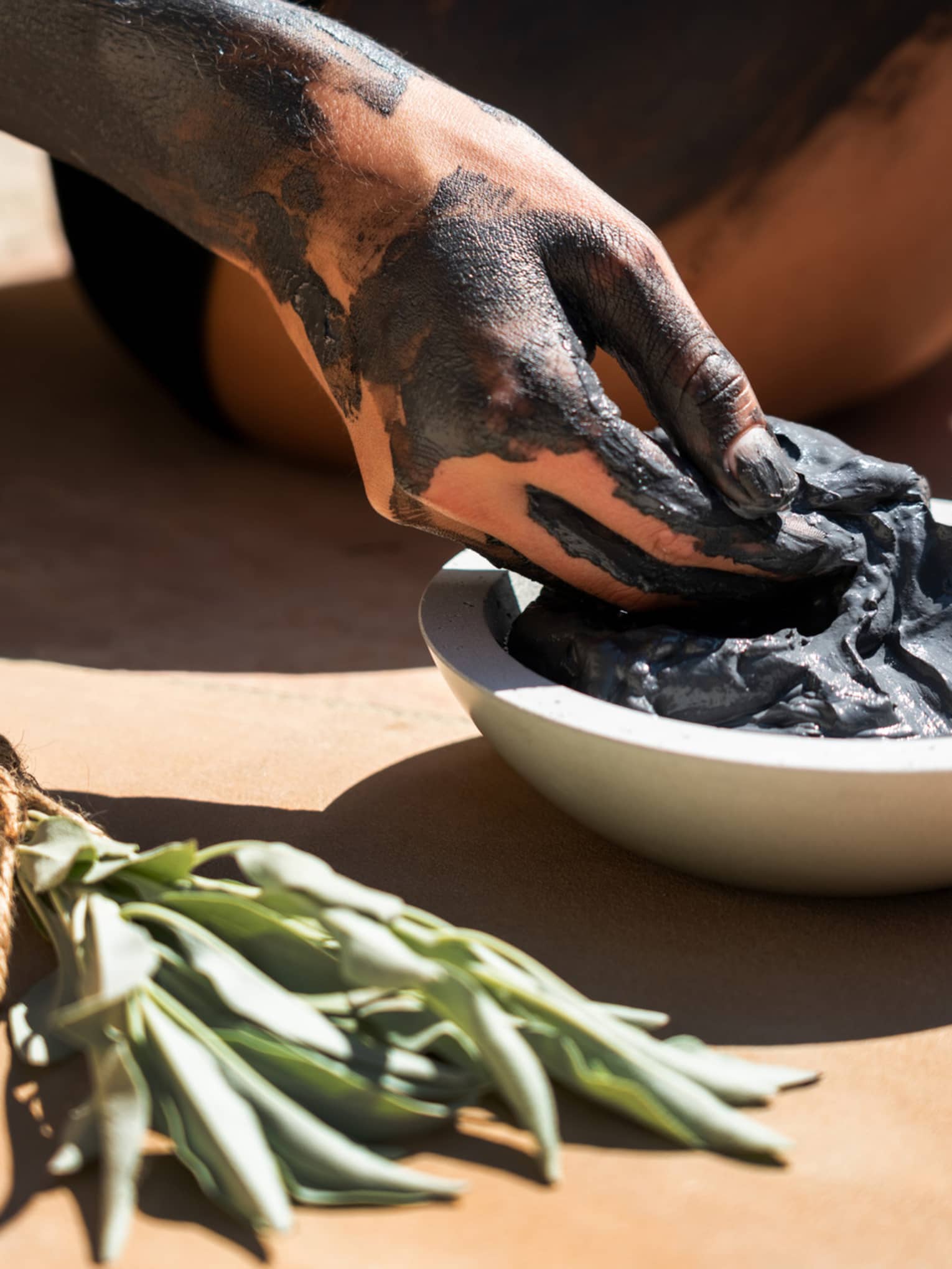 Hand covered in black clay reaching into a bowl of spa mud next to sage bundle