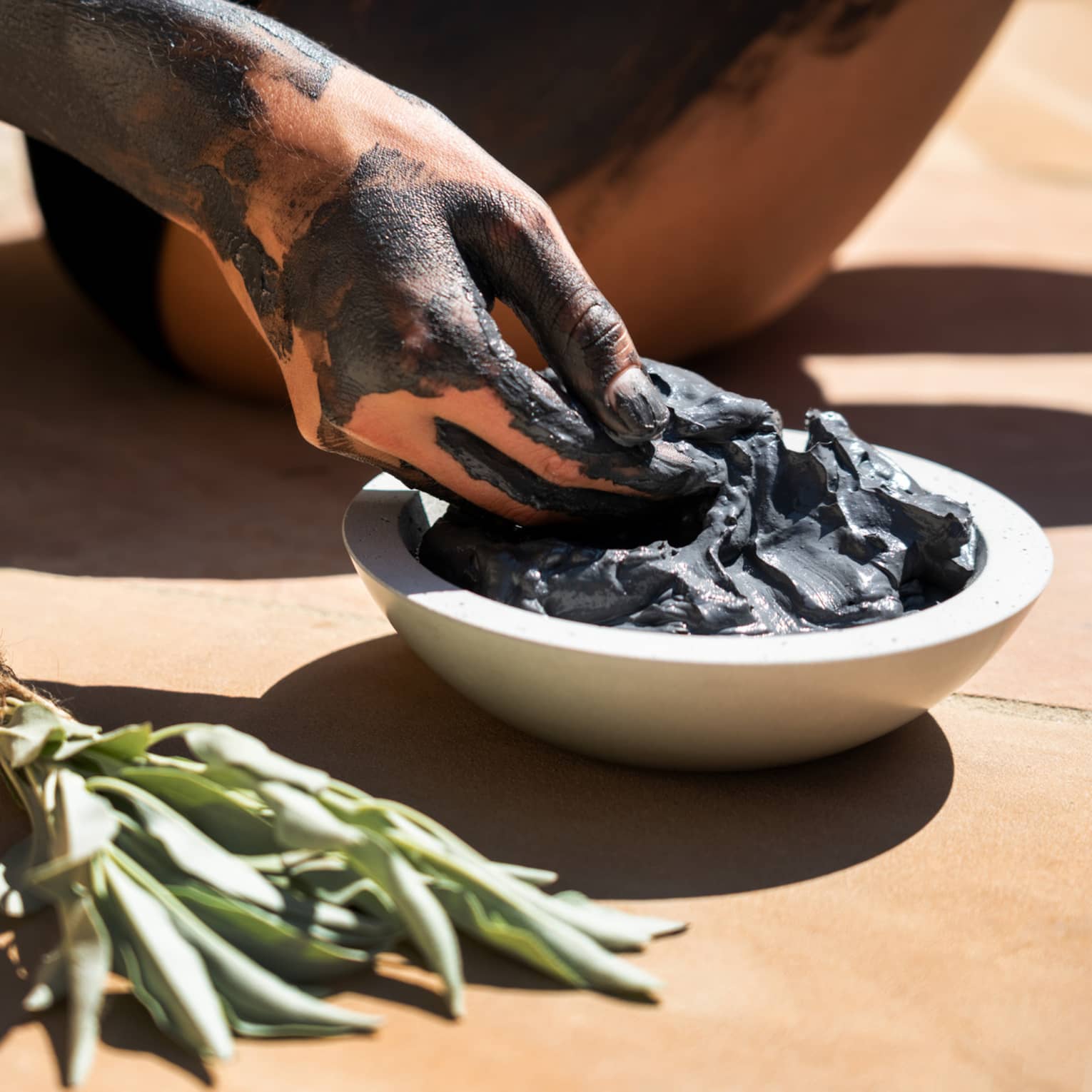 Hand covered in black clay reaching into a bowl of spa mud next to sage bundle
