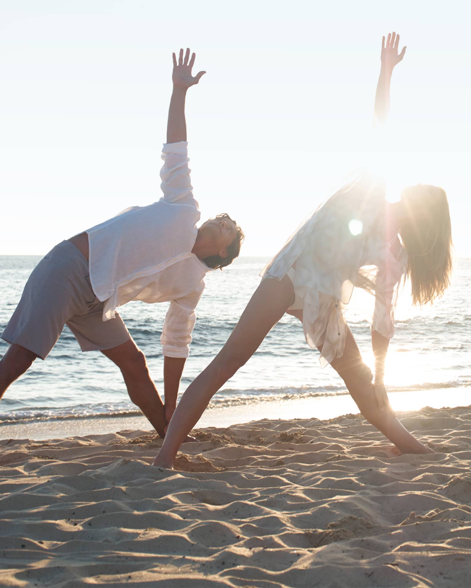 Two people in white beach shirts and shorts do a yoga pose with yogi at sunrise on beach