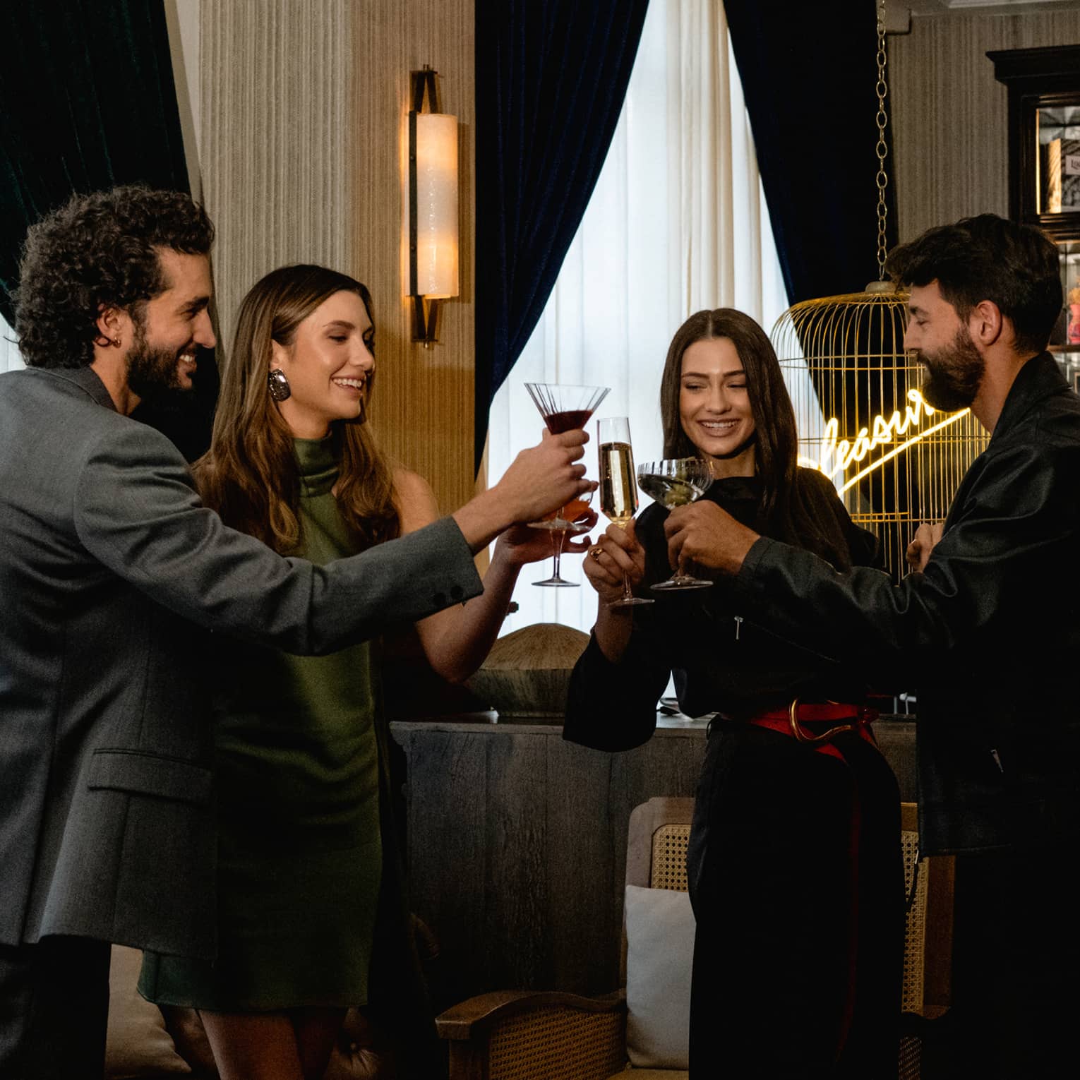 Four people raise their glasses in a toast in a cozy, softly lit lounge setting.