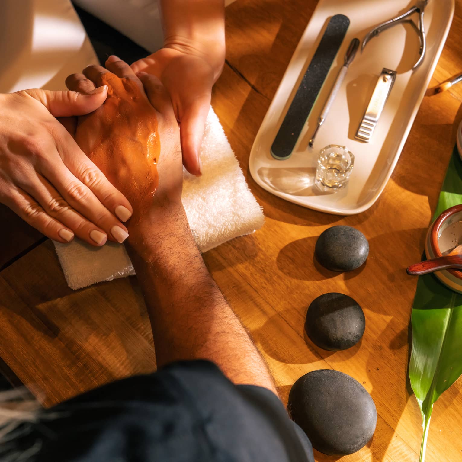 Manicurist massages a clients hand with three black stones, two small bowls of ingredients and nail tools sitting on the table