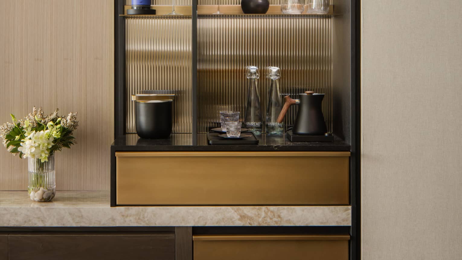 Stylish minibar with glass shelves displaying wine, glasses, and decorative items, next to a TV and vase with fresh flowers