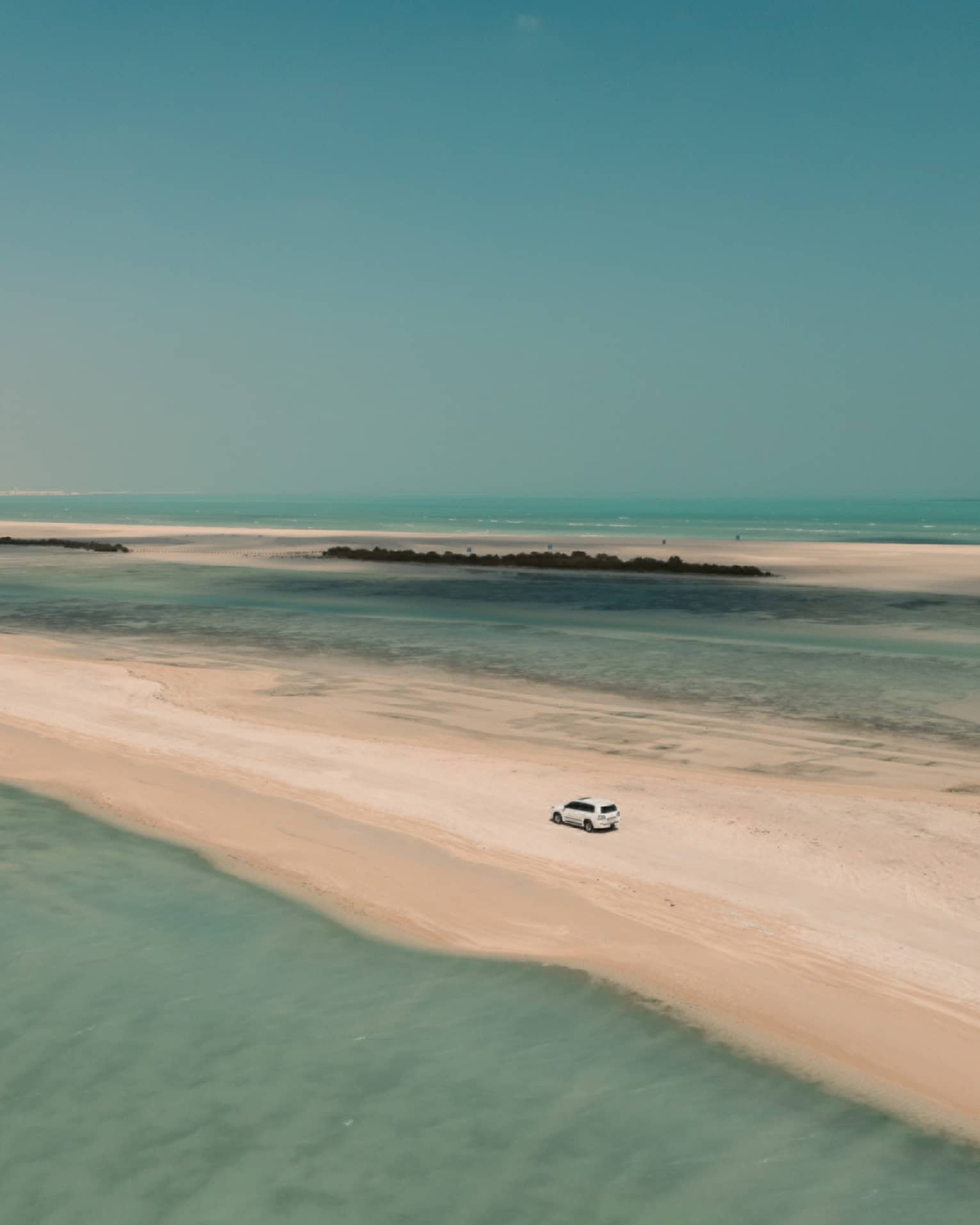 Aerial view of a 4x4 car traversing a sandbar surrounded by shallow turquoise water on either side and a cloudless sky above.