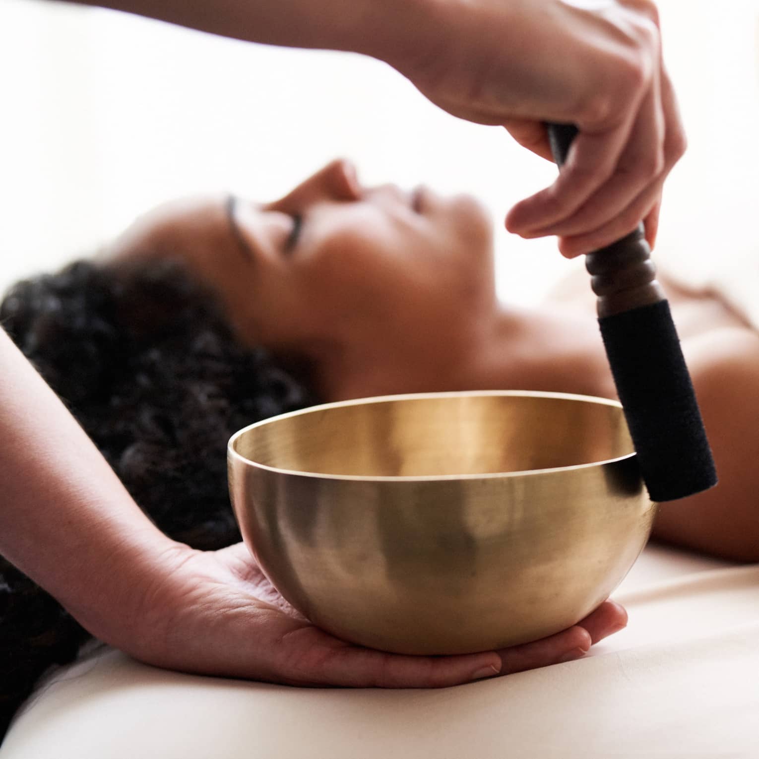 Woman with eyes closed lies on massage table in as vibrational sound therapy is performed with a Tibetan singing bowl