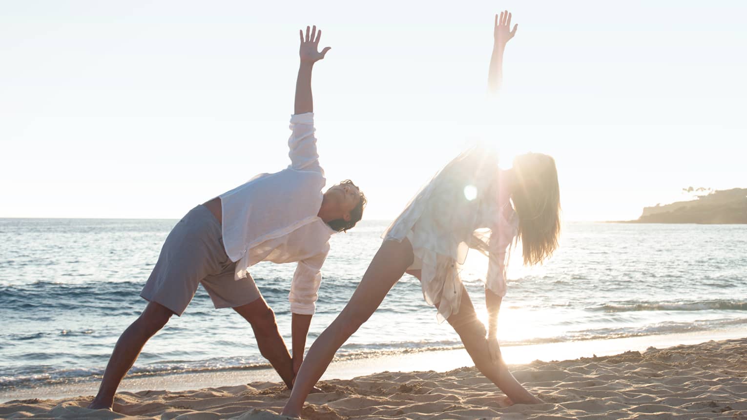 Two people in white beach shirts and shorts do a yoga pose with yogi at sunrise on beach
