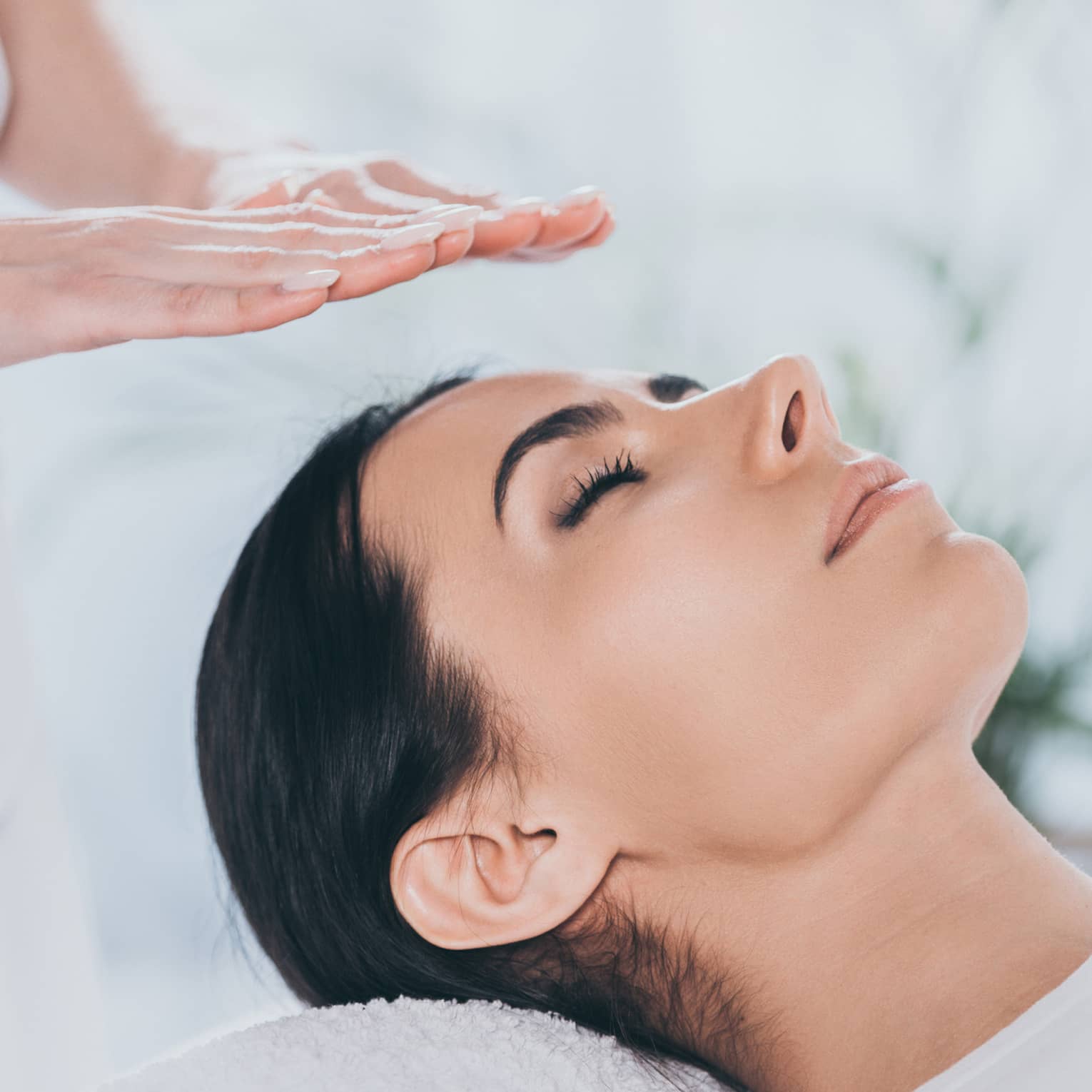 Woman with eyes closed lays on rolled up towel on spa table under therapists' hovering hands