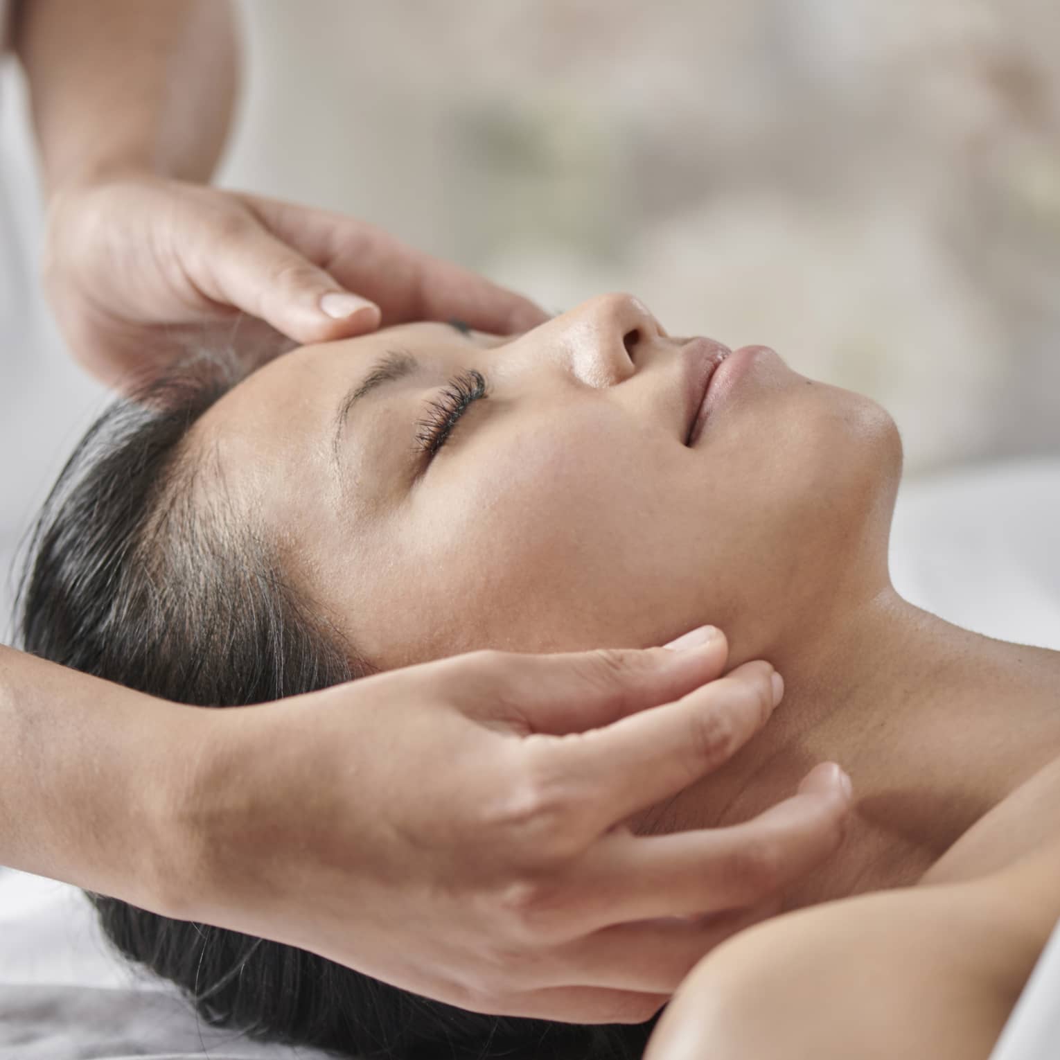 Masseuse massages woman's face as she lies face up on table under white sheet