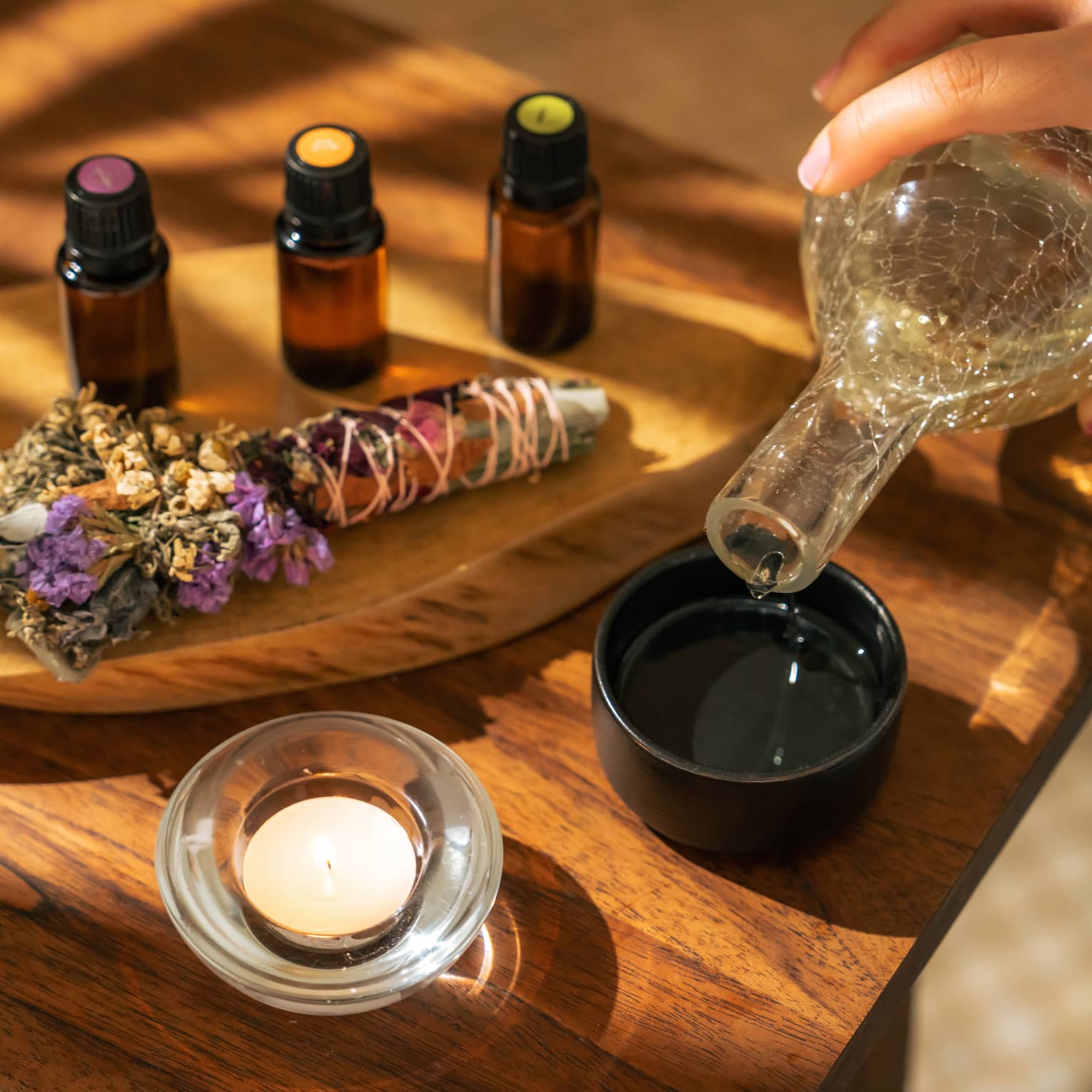 Four small bottles of essential oils and a sage-and-floral bundle stick lie on a wooden platter next to a hand pouring liquid into a small black bowl next to a votive candle
