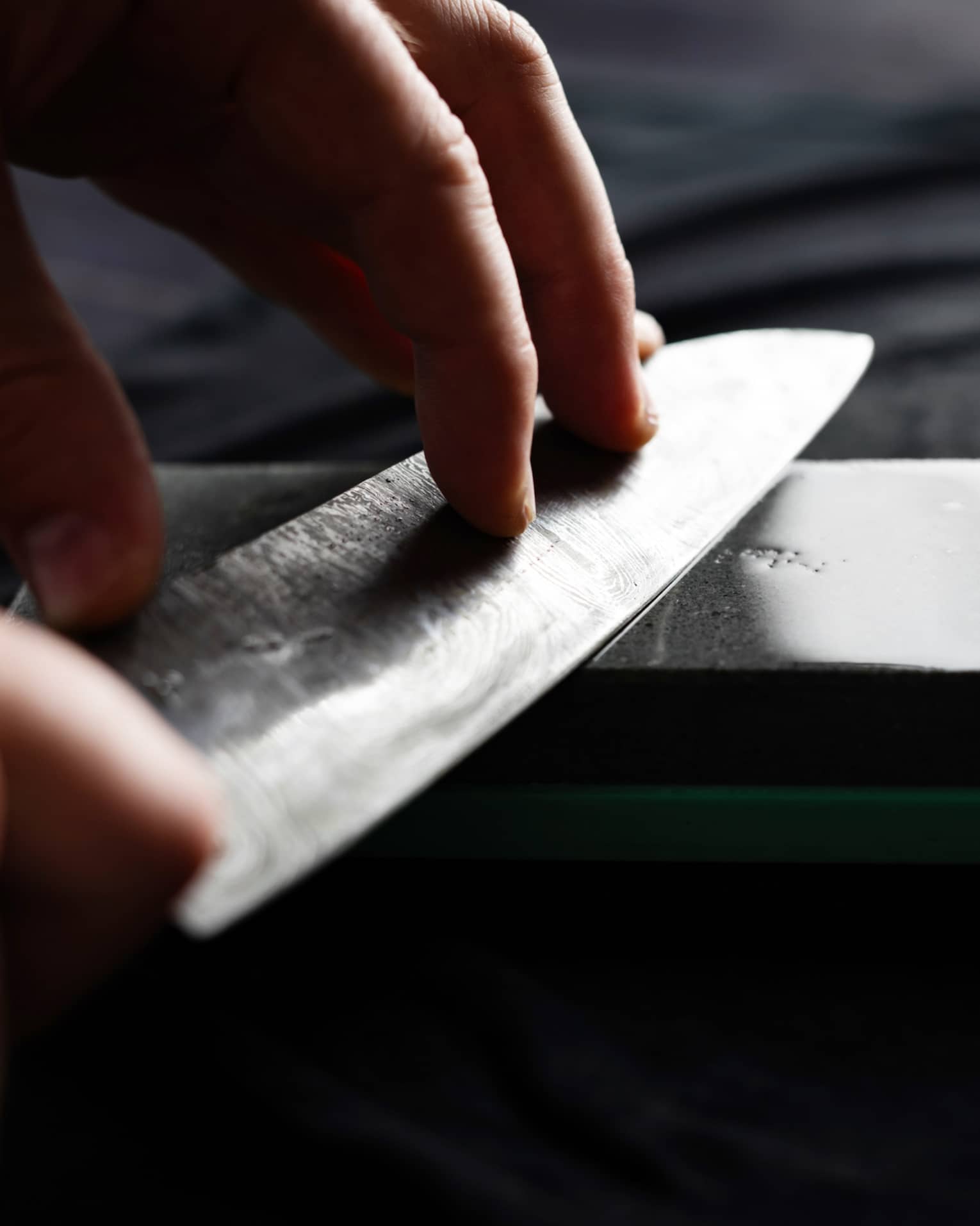 Close-up of two hands holding a chef’s knife against a gleaming dark-grey sharpening stone on a dark surface.