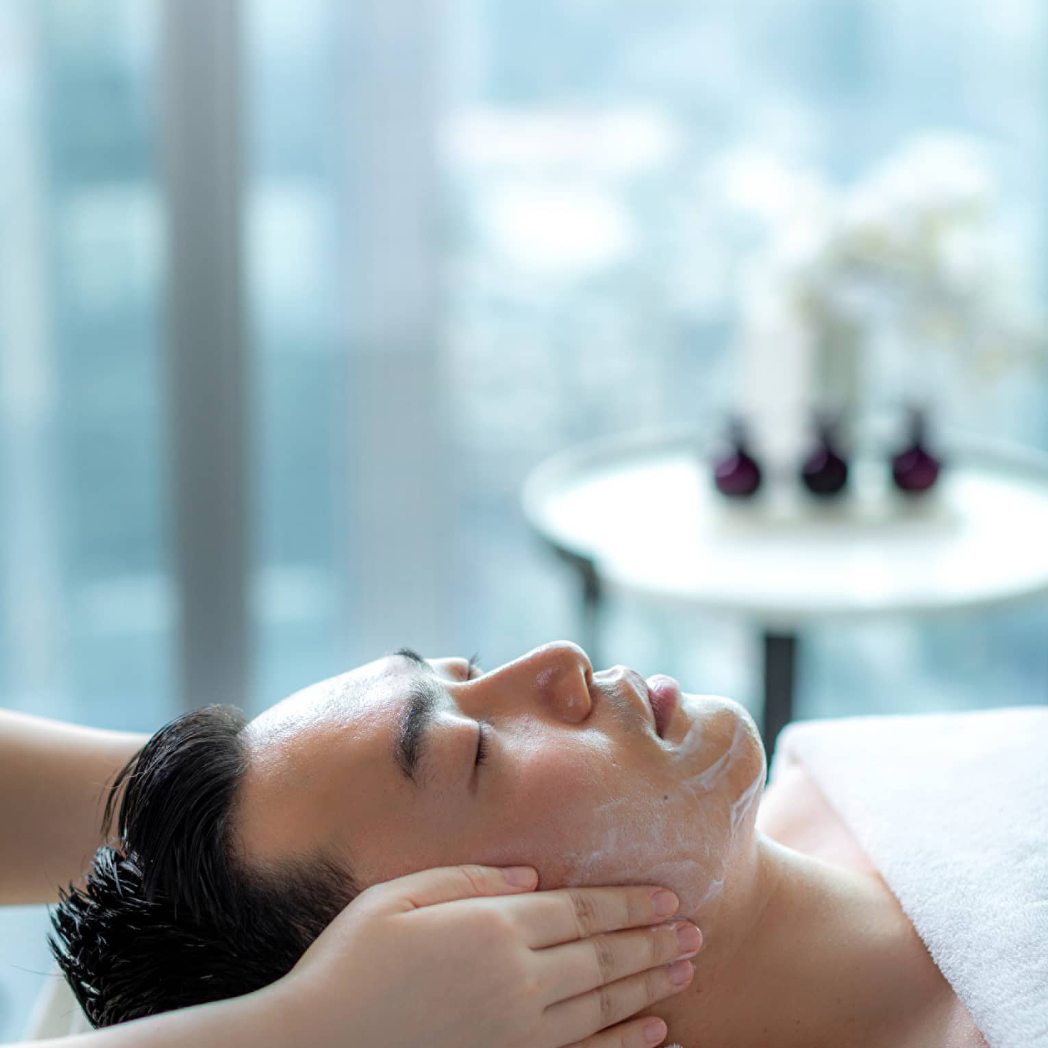 Hands rest on man's cheeks as he lays on massage table by glass window with daytime city view