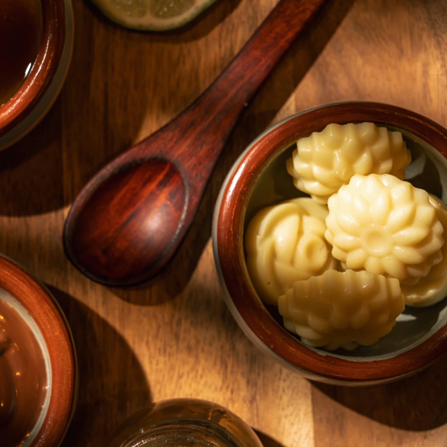 Three stoneware bowls filled with spa ingredients and a wooden spoon sit on a round wooden table