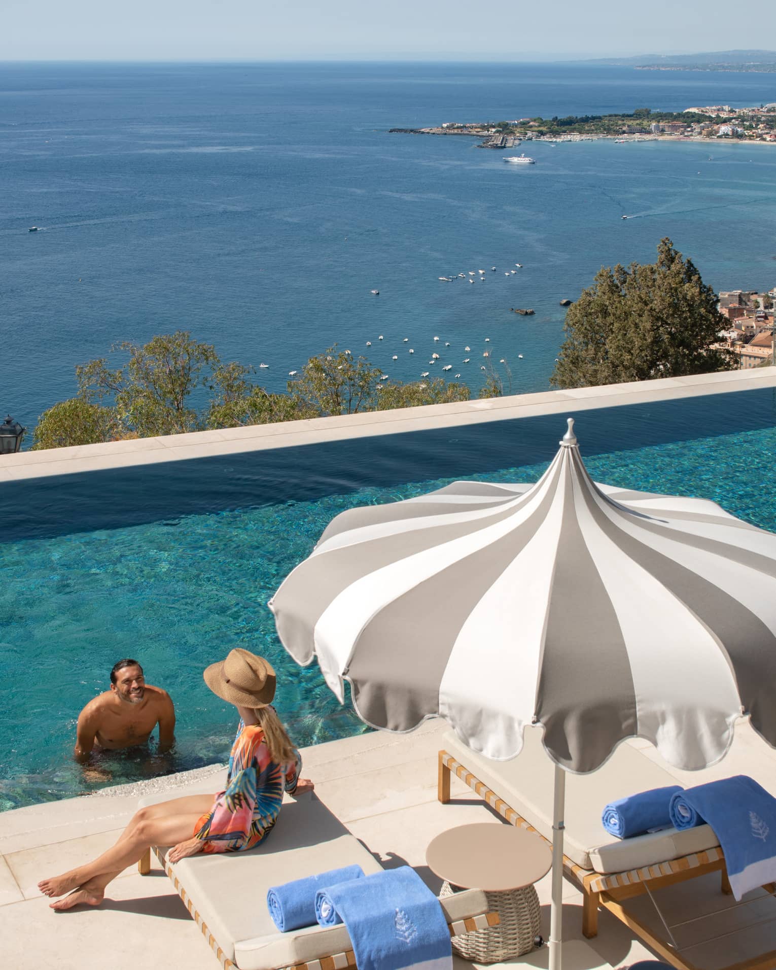 A man at the edge of the infinity pool speaking to a woman sitting on a deck chair