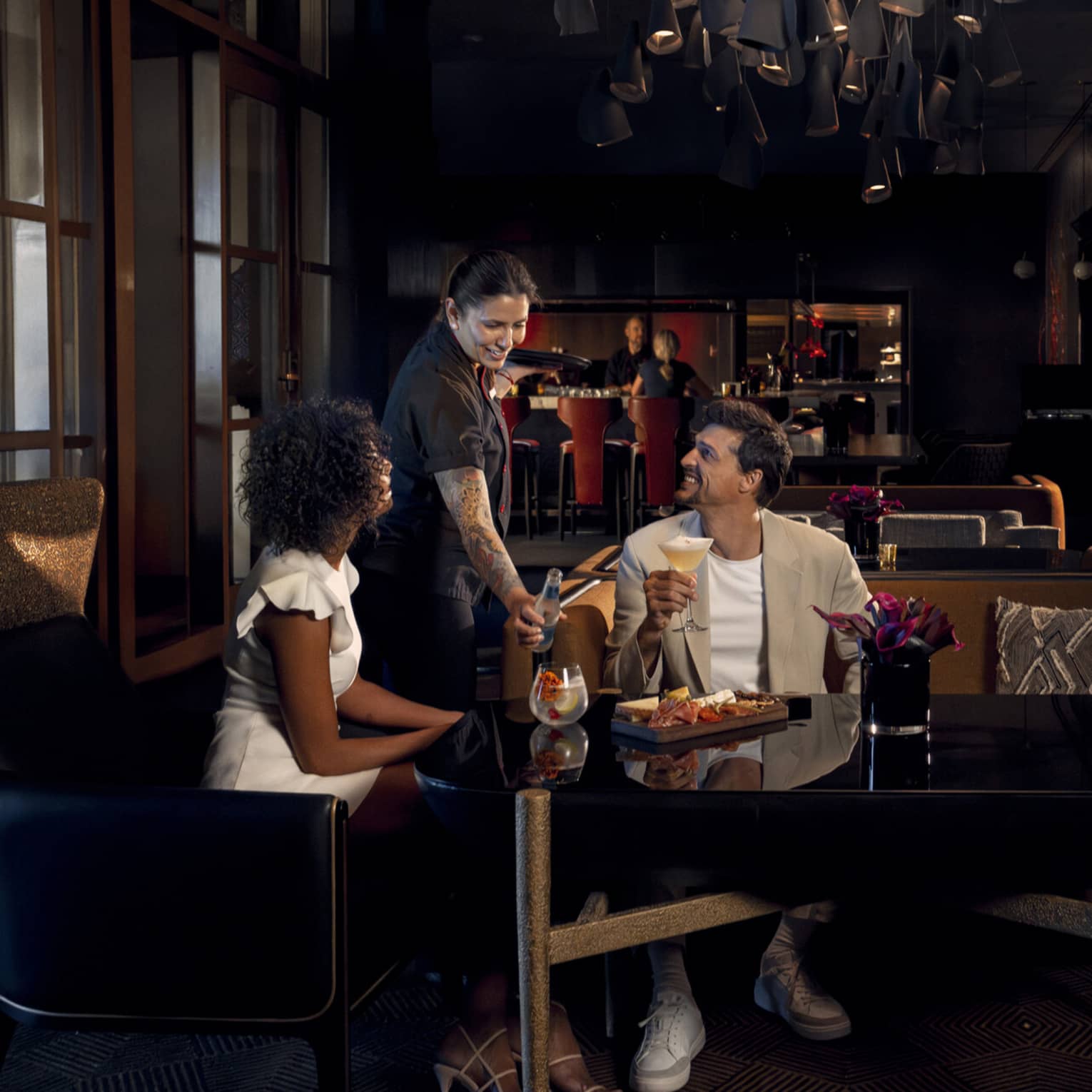 Two people wearing light-coloured cocktail attire sit on benches around a black table as a waitress brings them beverages