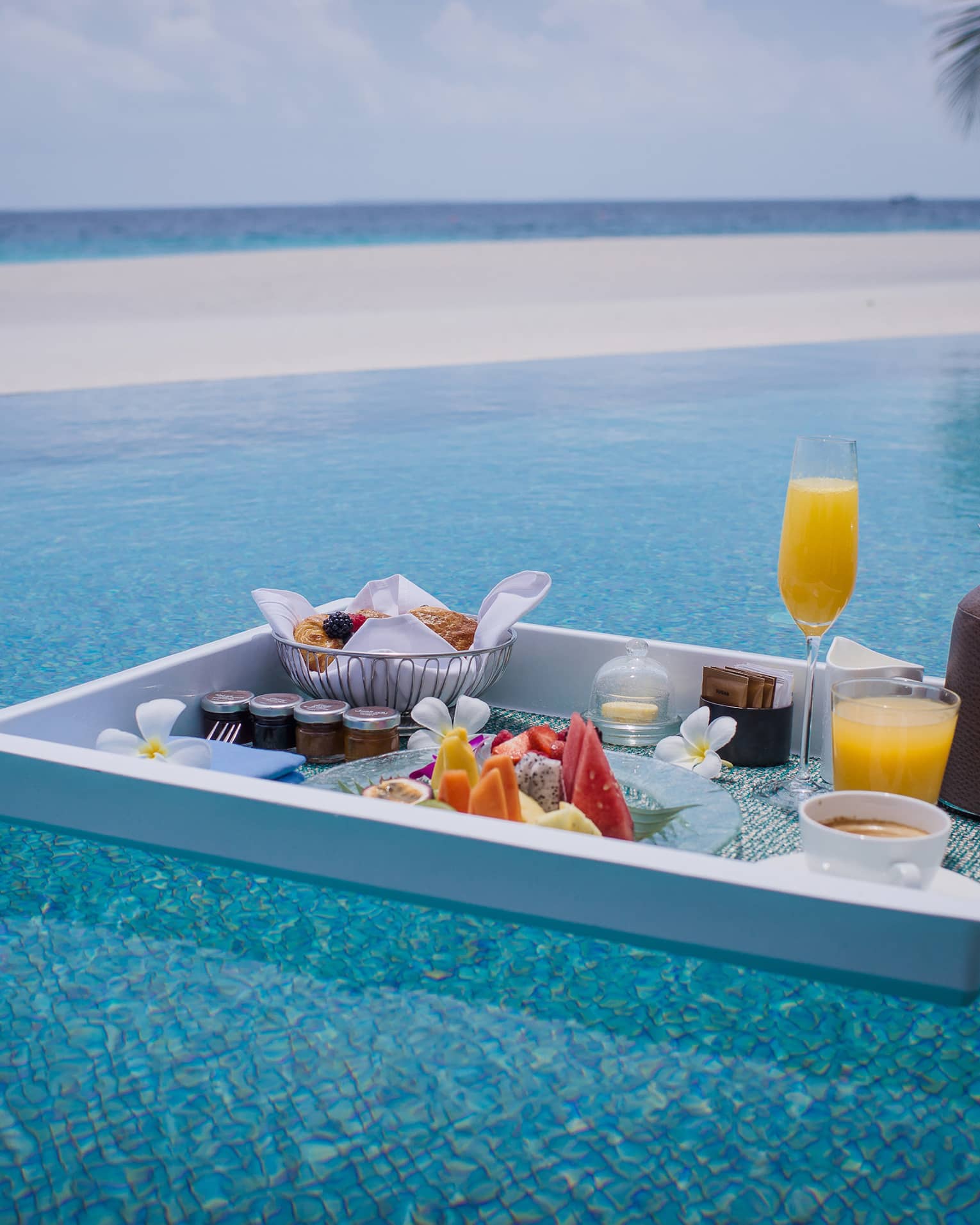 Floating in a shimmering turquoise pool is a white tray with a selection of fruit, pastries, jams, beverages and flowers.
