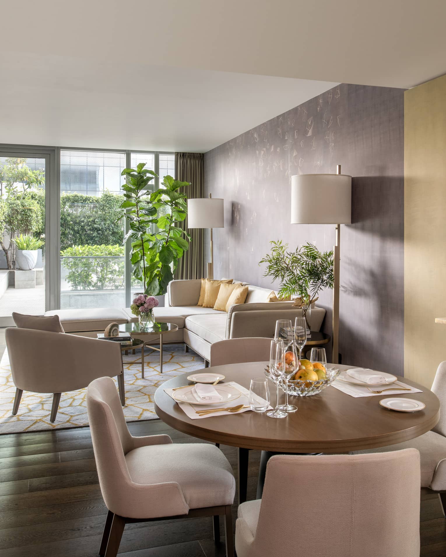 Guest room living and dining area with cream-coloured sectional sofa, round wooden dining table with four chairs, gold accents, floor-to-ceiling sliding glass door to patio