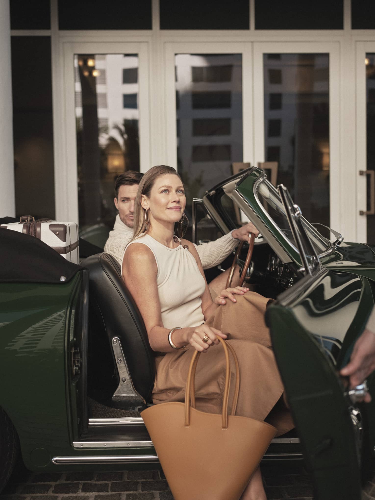 A man and woman getting out of a dark green car holding bags, two hotel team members are assisting them.