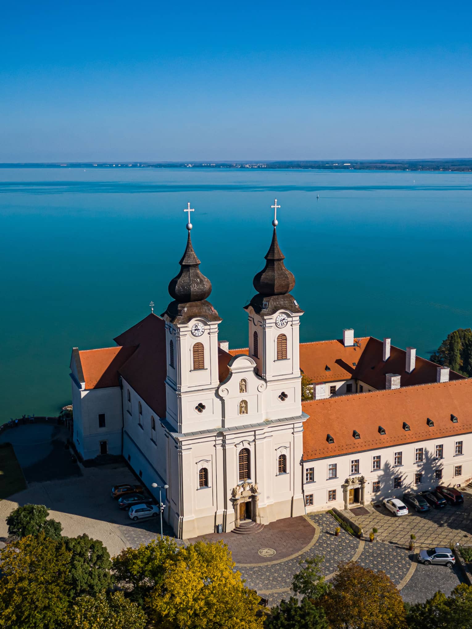 A Benedictine monastery, with white walls and a red tiled roof, sitting beside a large blue lake and surrounded by trees.