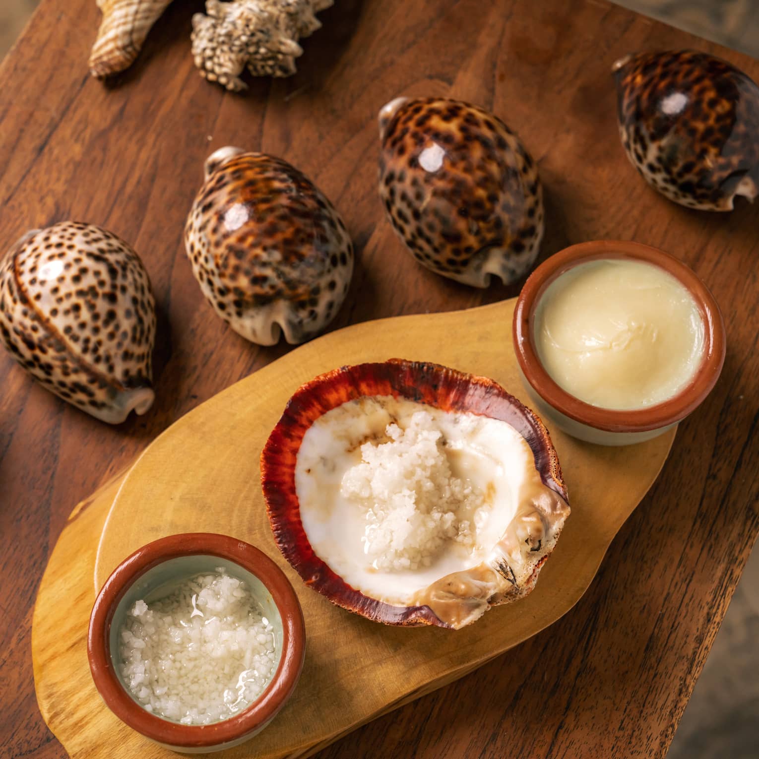 Five cowrie shells lined up in a row on a wooden table sit next to three small red bowls of body scrubs