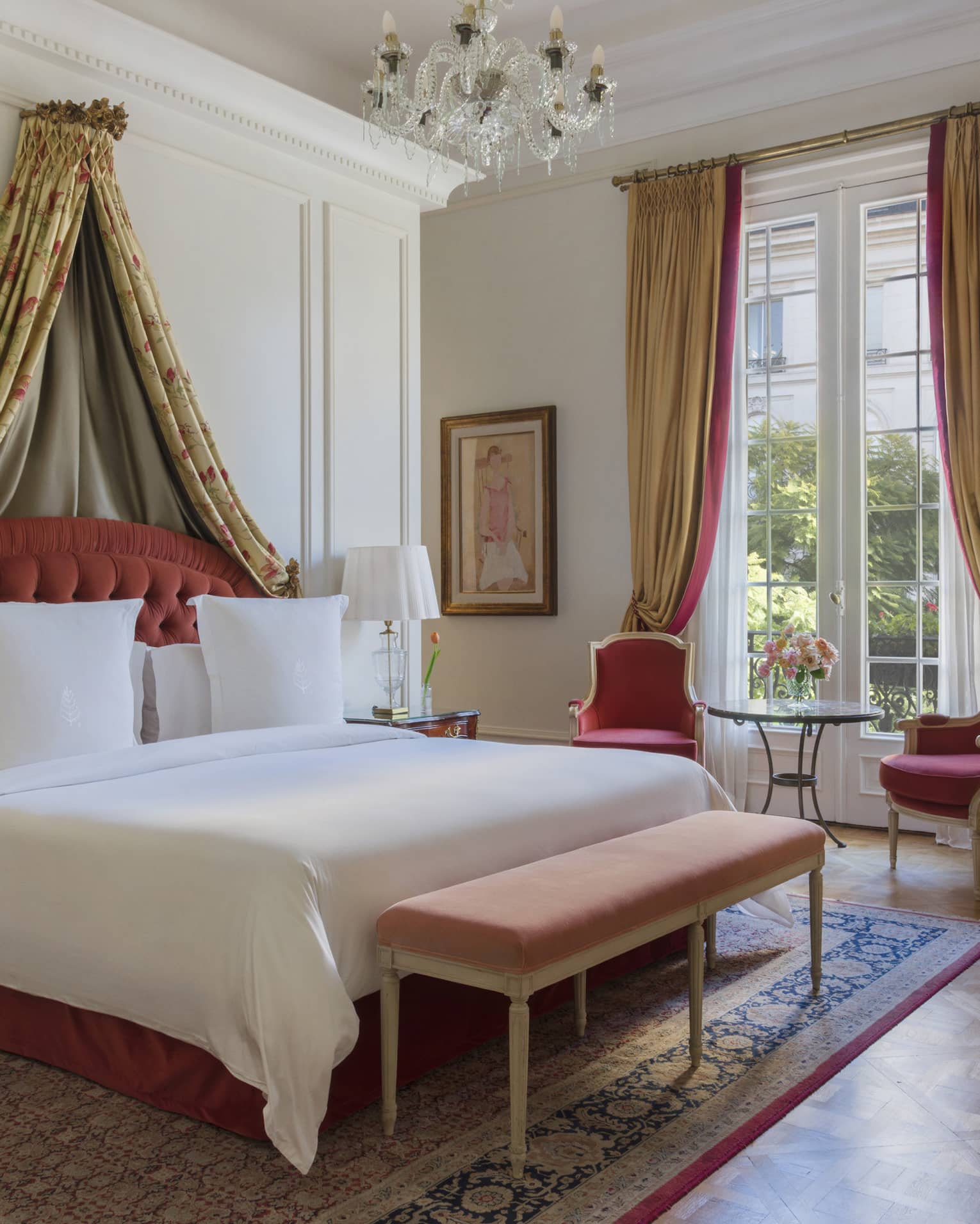 Lavish hotel suite bedroom, featuring a king bed with draped canopy and red armchairs by a window