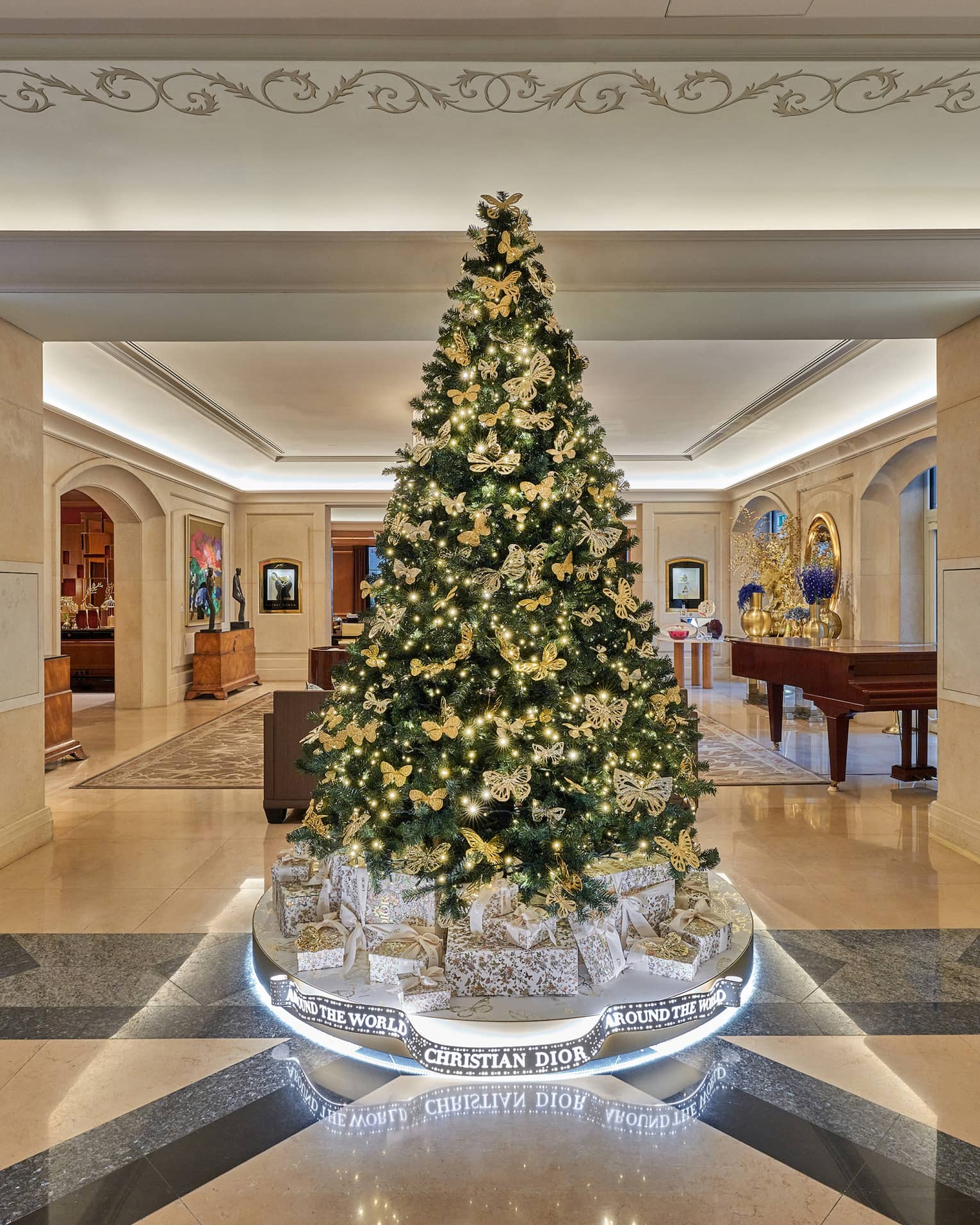 Luxurious hotel lobby, featuring a large elegantly decorated Christmas tree with gold ornaments and lights, surrounded by Christian Dior gift boxes and modern artwork on the walls.