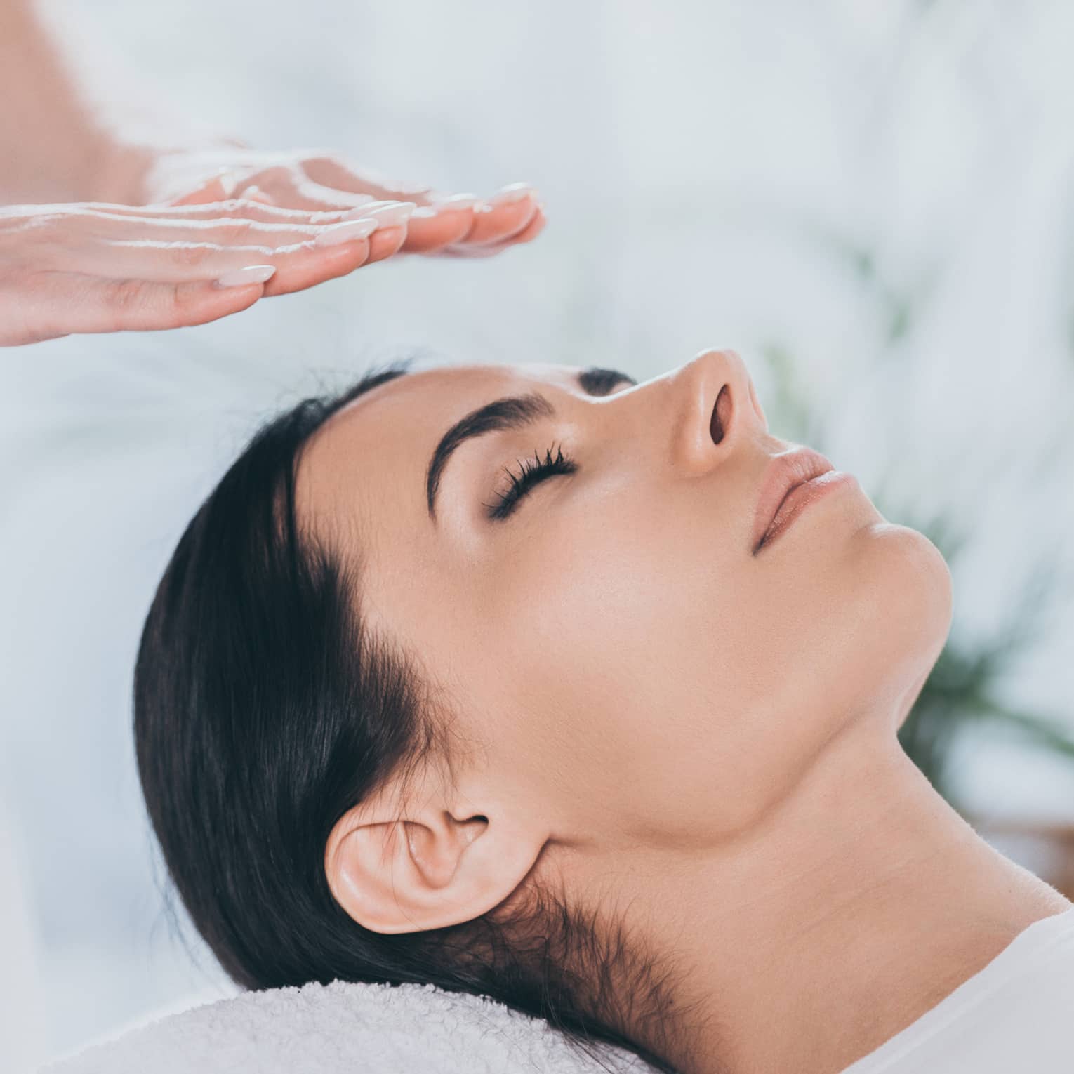 Woman with eyes closed lays on rolled up towel on spa table under therapists' hovering hands