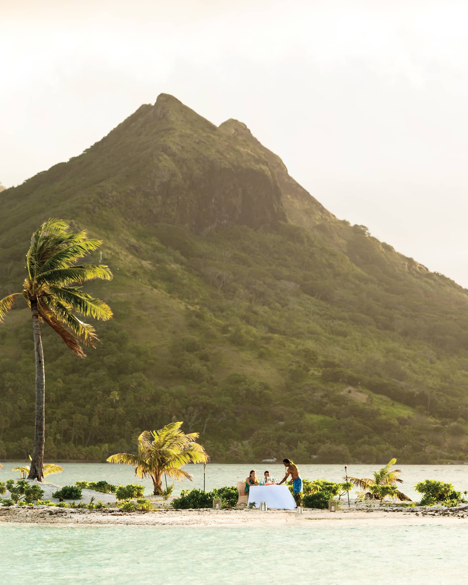 Small tropical island with palm trees, where two people are dining at a table by the water, with a mountain in the background