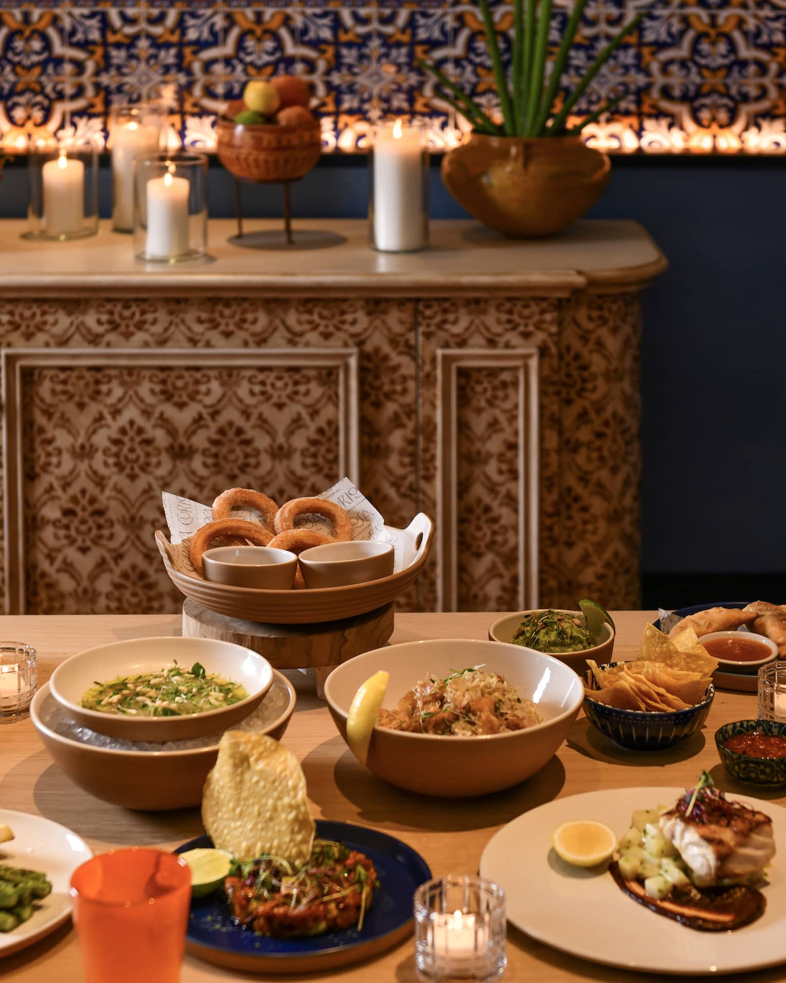 A table set with a variety of appetizers on white plates and bowls with a sideboard in the background adorned with white pillar candles