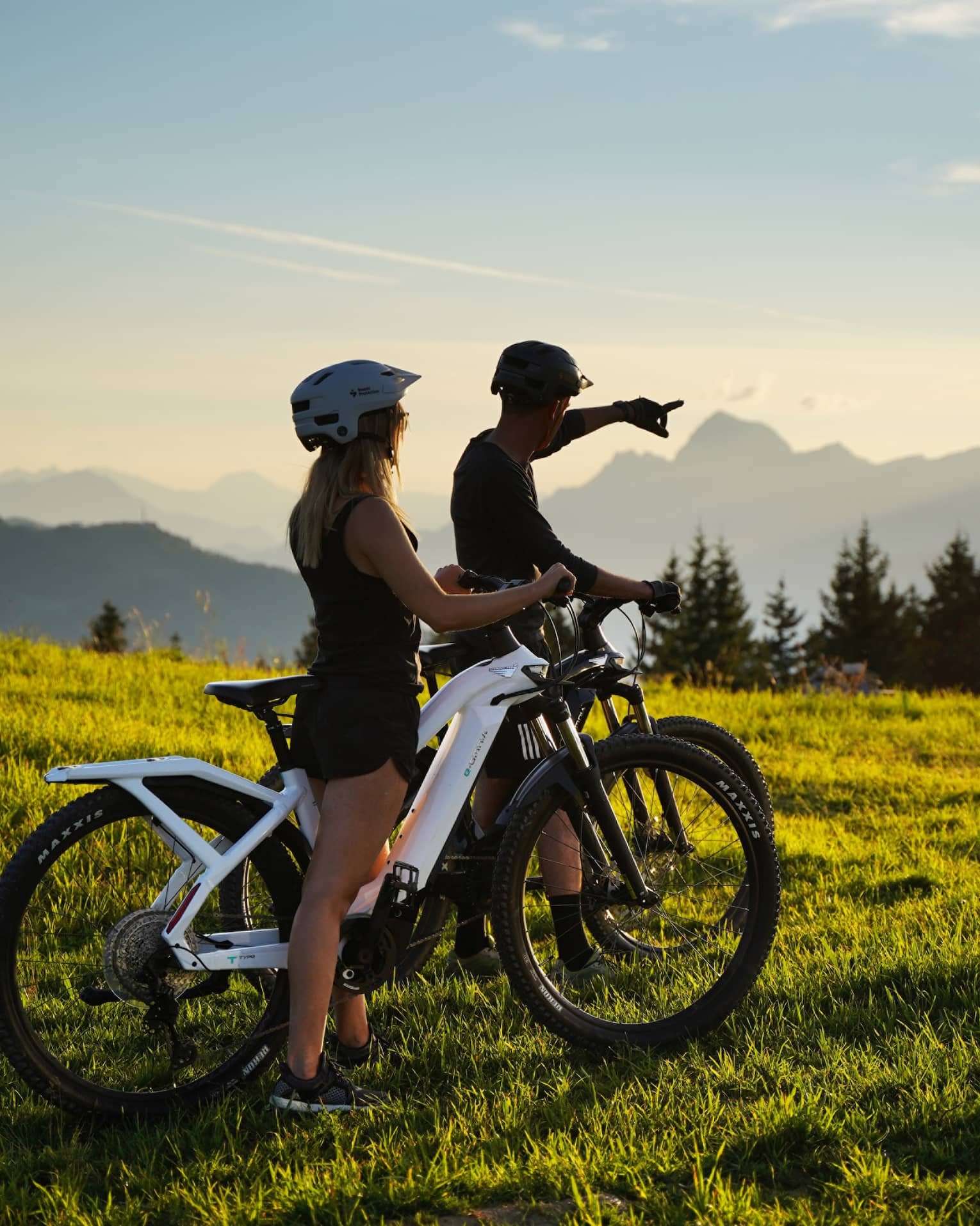 Two people riding fat bikes on meadow, one pointing to distant mountains