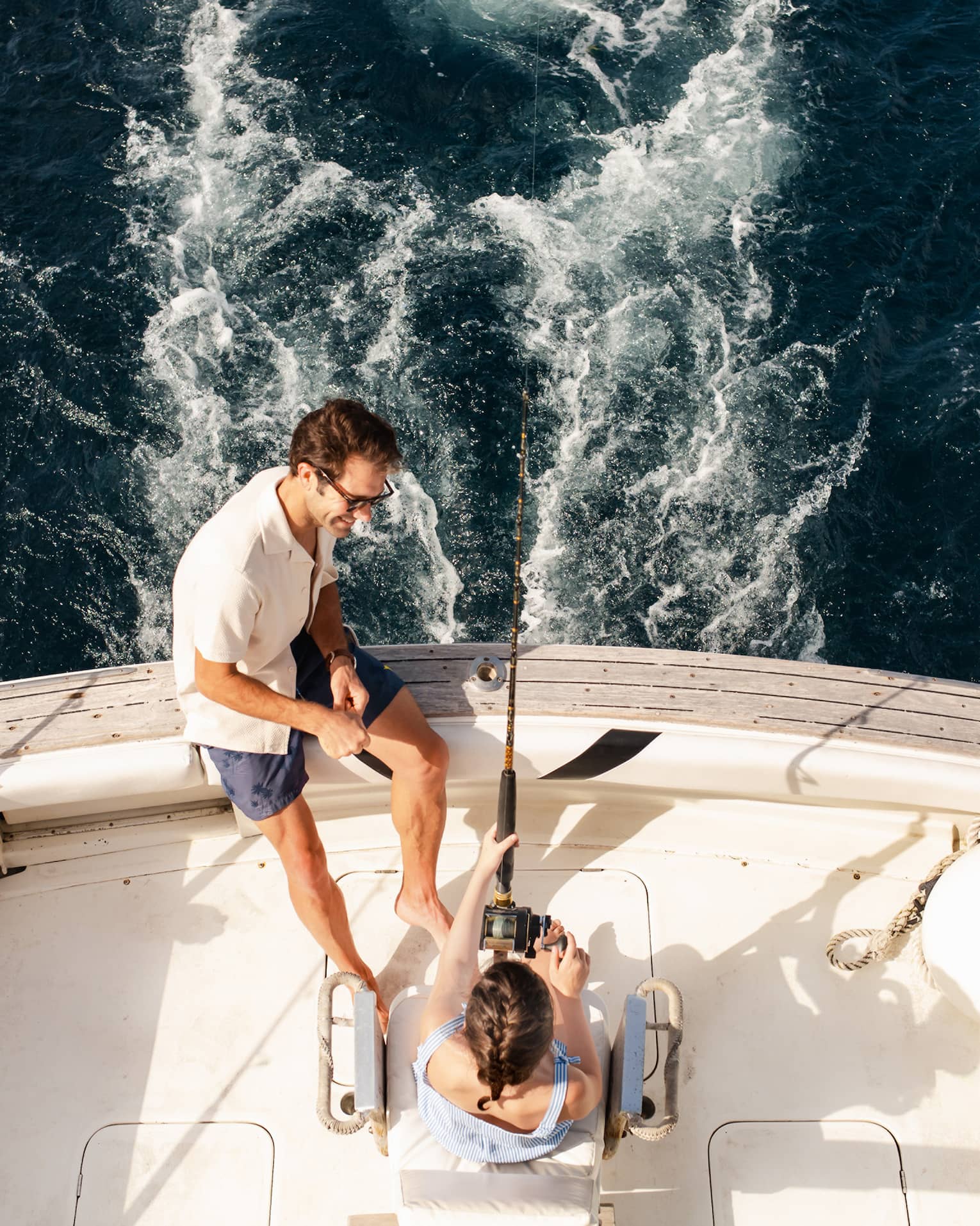 Two people sit at the back of a fishing boat, with one person holding a fishing rod and the other sitting close by on the edge of the boat