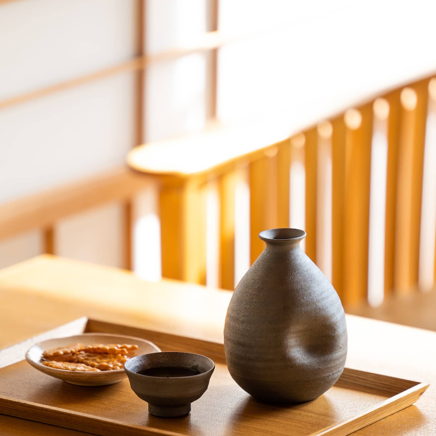 Close-up of a dark ceramic sake carafe with matching cup and a small plate of crackers on a wooden tray and table.