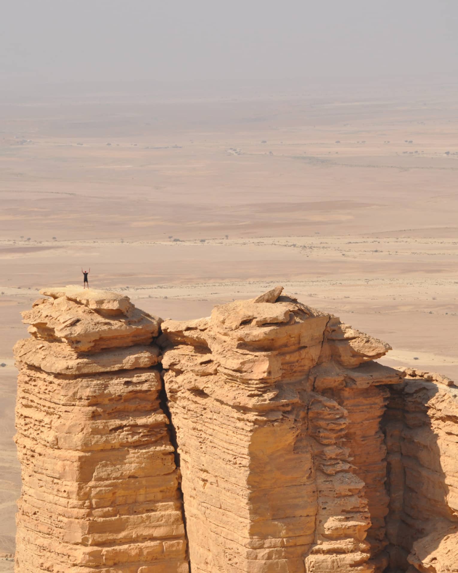 An aerial view of a giant golden cliff surrounded by a dry desert and a small silhouette of a person standing on top of it.