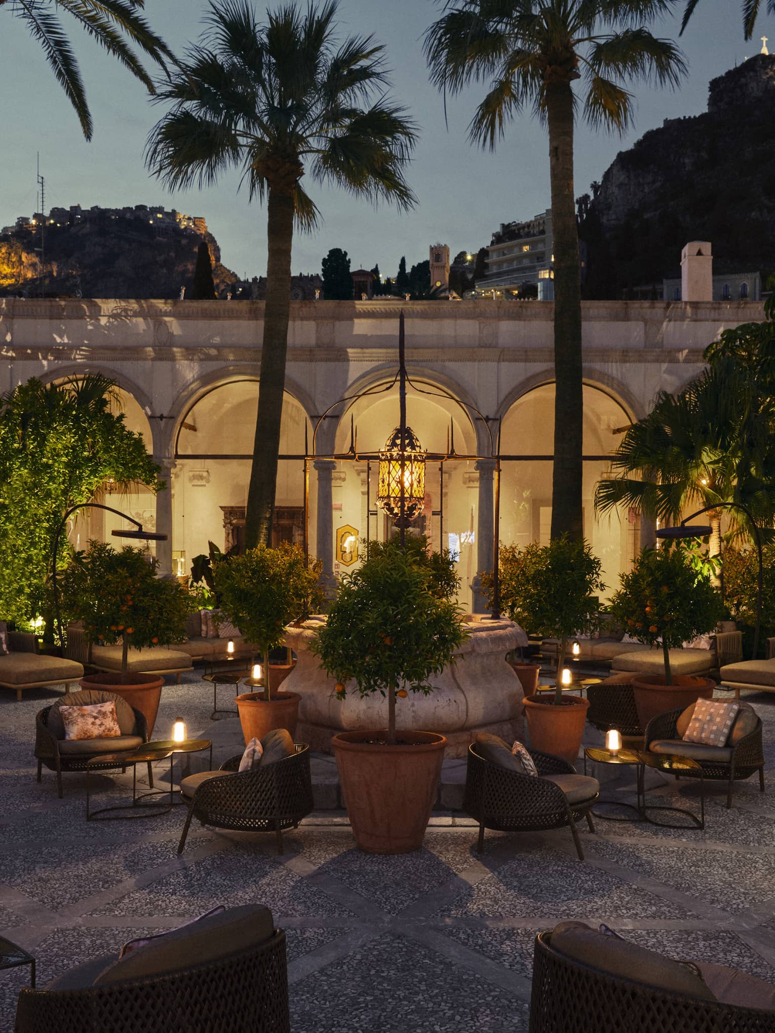 Courtyard-style lounge area with various seating areas and tables topped with candles, all set beneath some trees with an arched colonnade walkway in the background at night