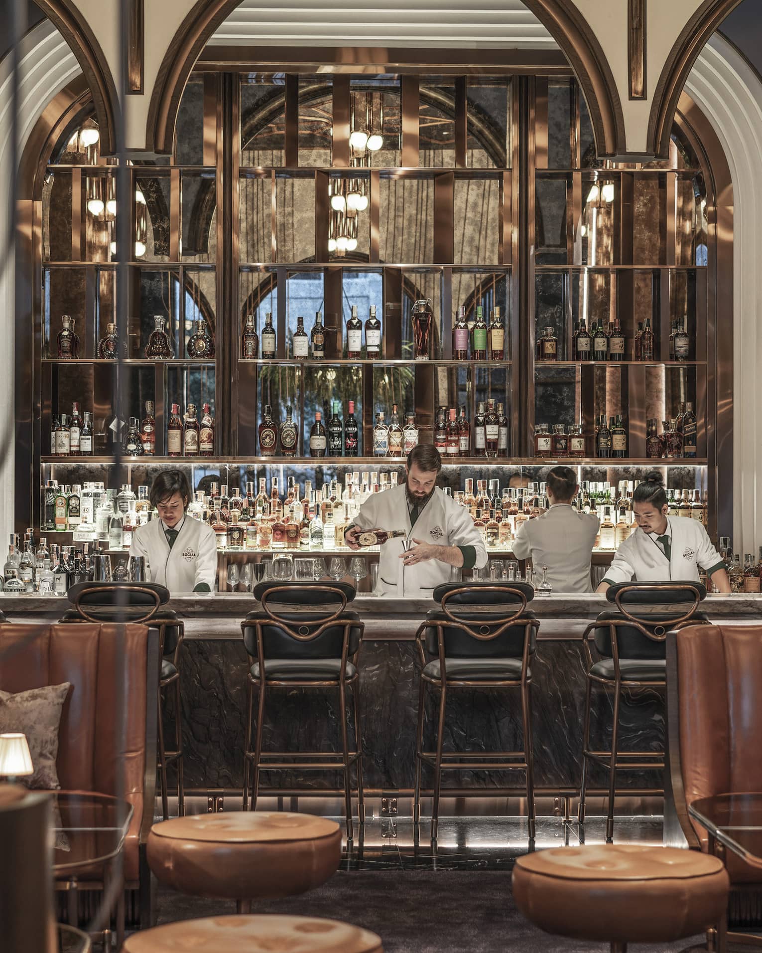 Chic bar with leather seating, plants on the walls and a well-stocked backlit bar, where bartenders in white jackets prepare drinks behind the counter.