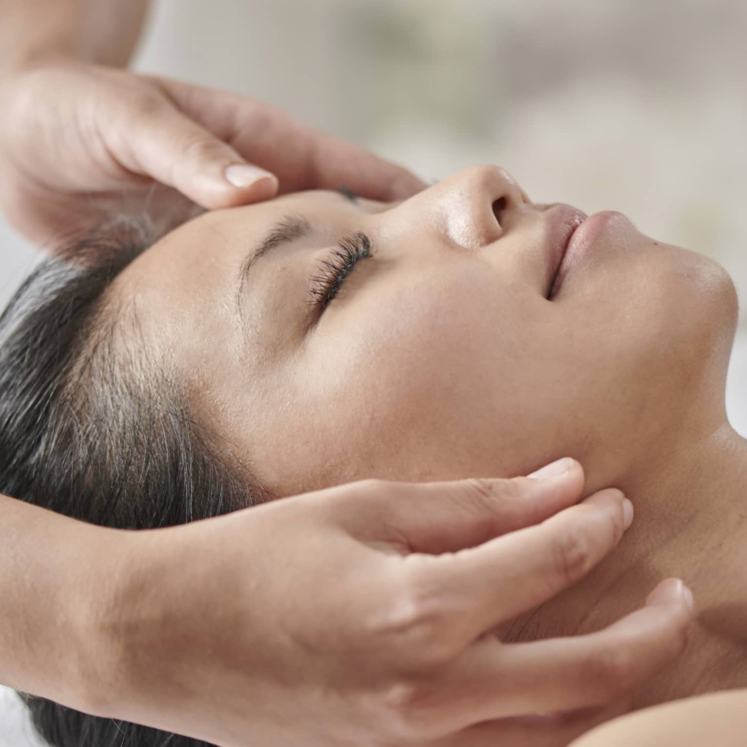 Masseuse massages woman's face as she lies face up on table under white sheet