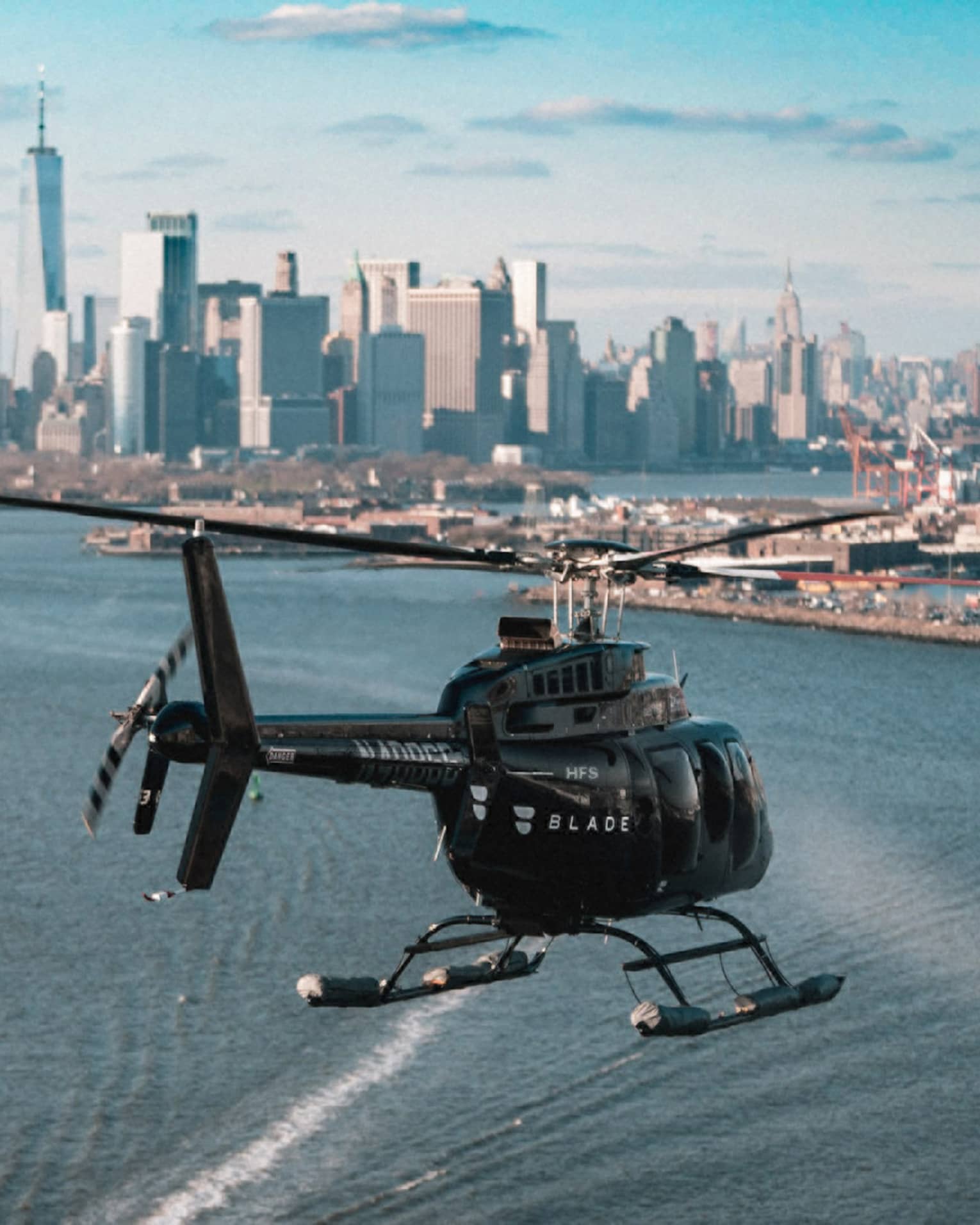 Rear view of a helicopter flying over water in the direction of a shipyard and the iconic New York skyline in the distance.