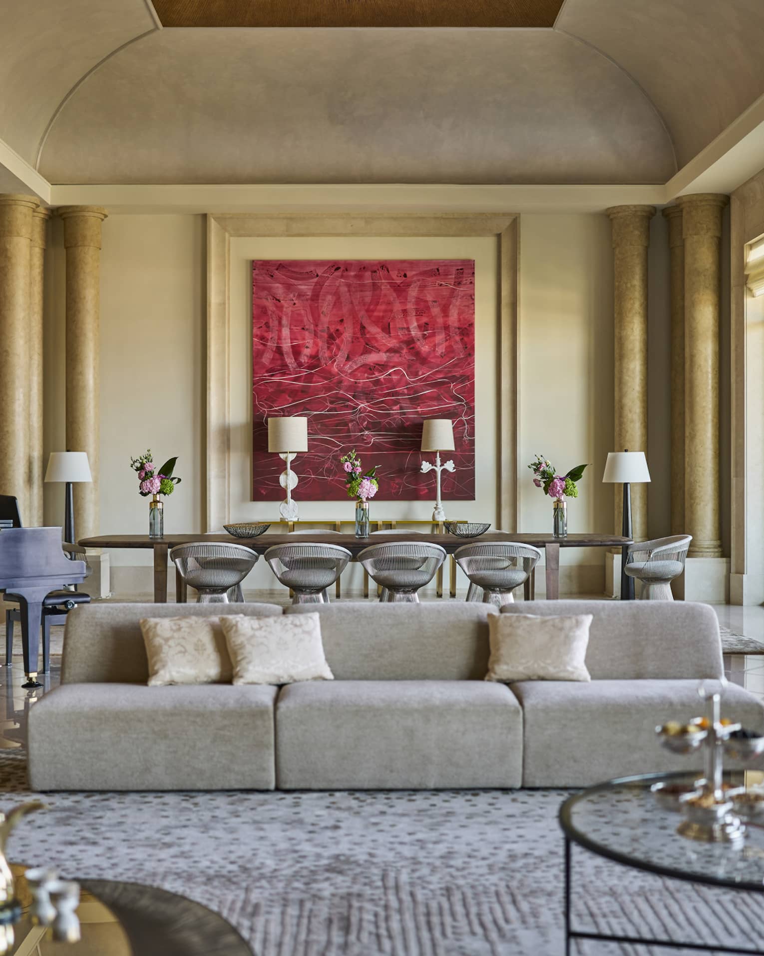 Grand hotel suite living room with pillars, a modern sofa and a large dining table set next to red artwork