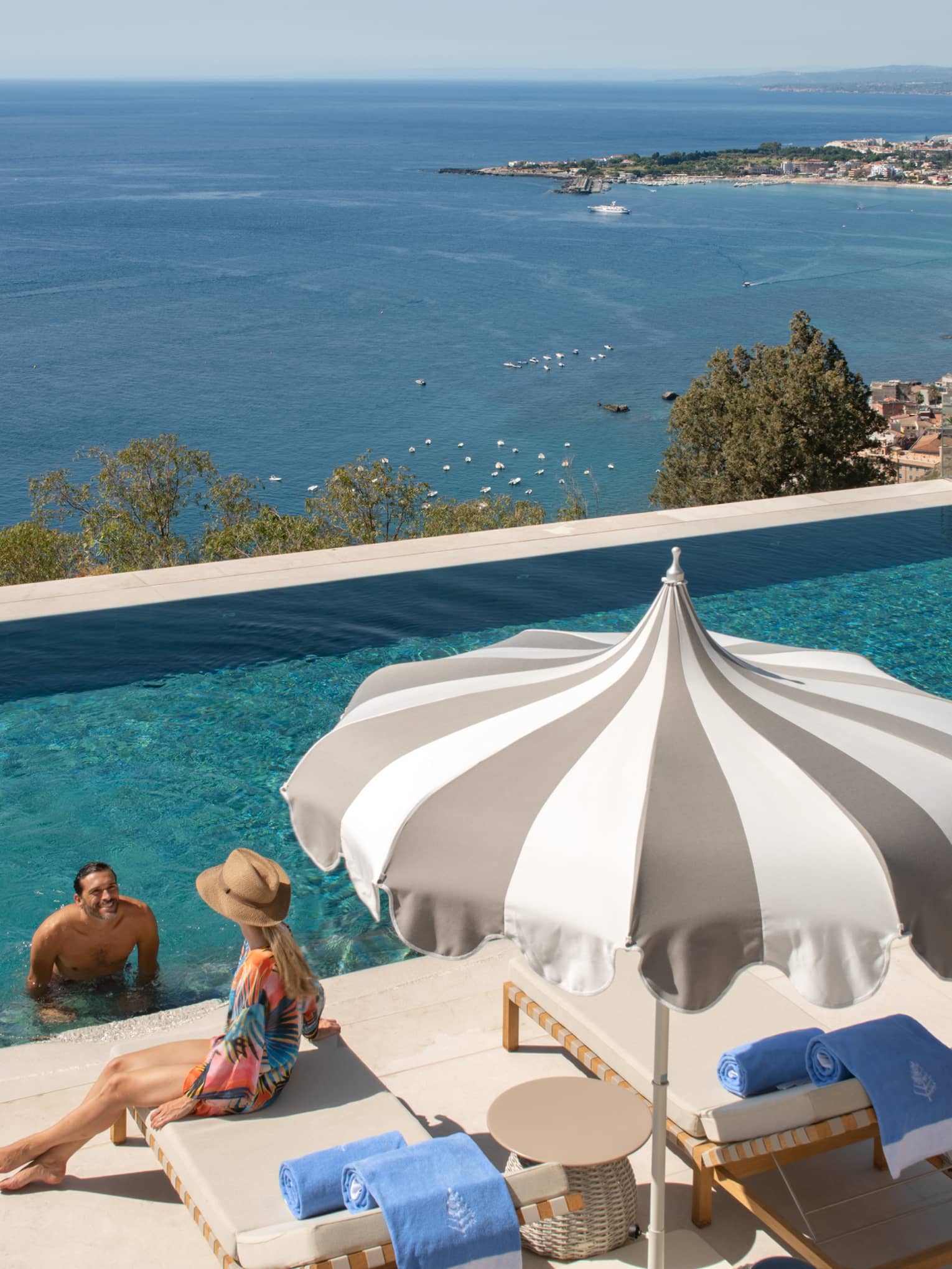 A man at the edge of the infinity pool speaking to a woman sitting on a deck chair